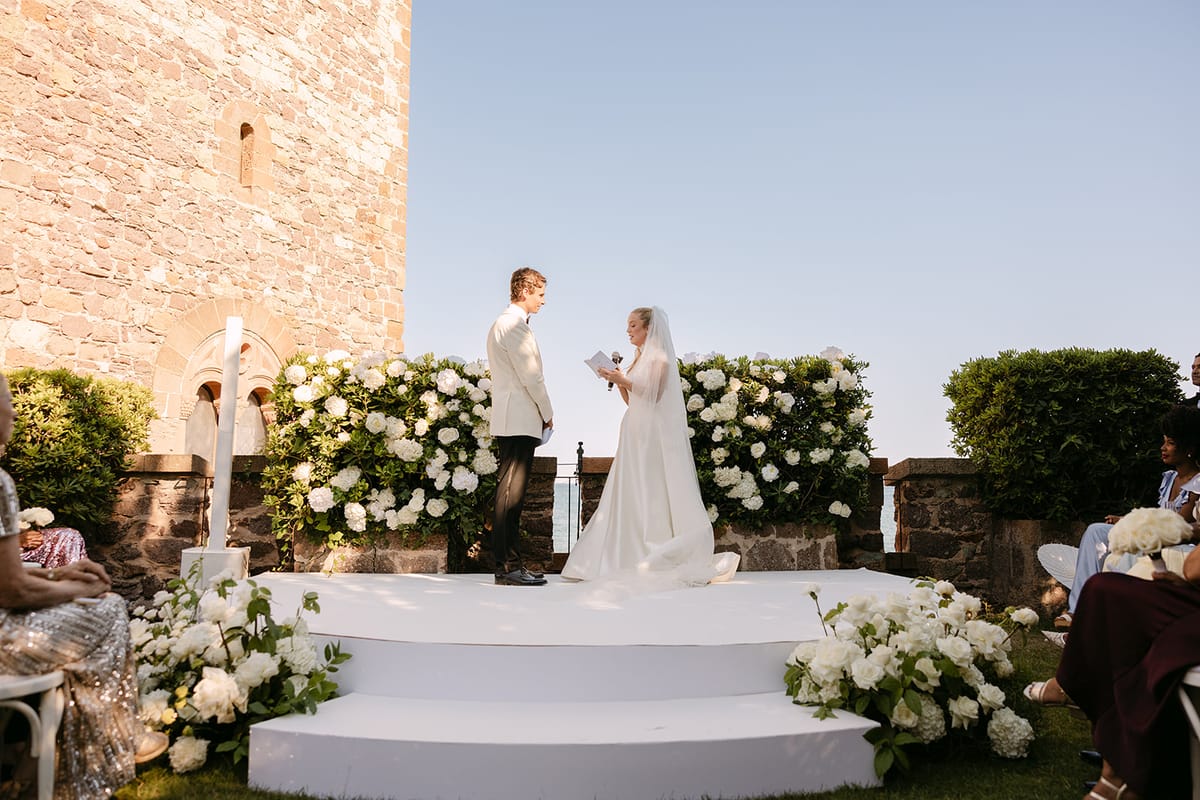 The couple stand facing each other on a white stepped platform surrounded by white hydrangeas, with a stone tower and clear sky behind.