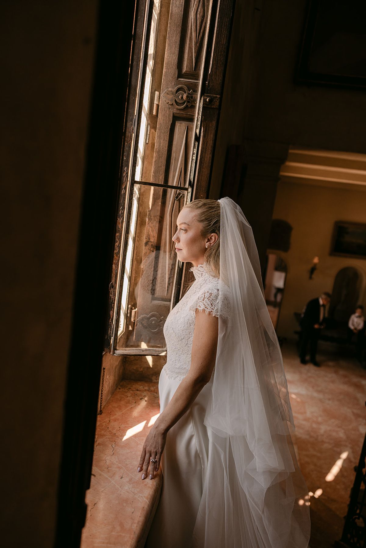 The bride, wearing a lace-sleeved dress and long veil, stands in profile beside a tall window in a dim room.