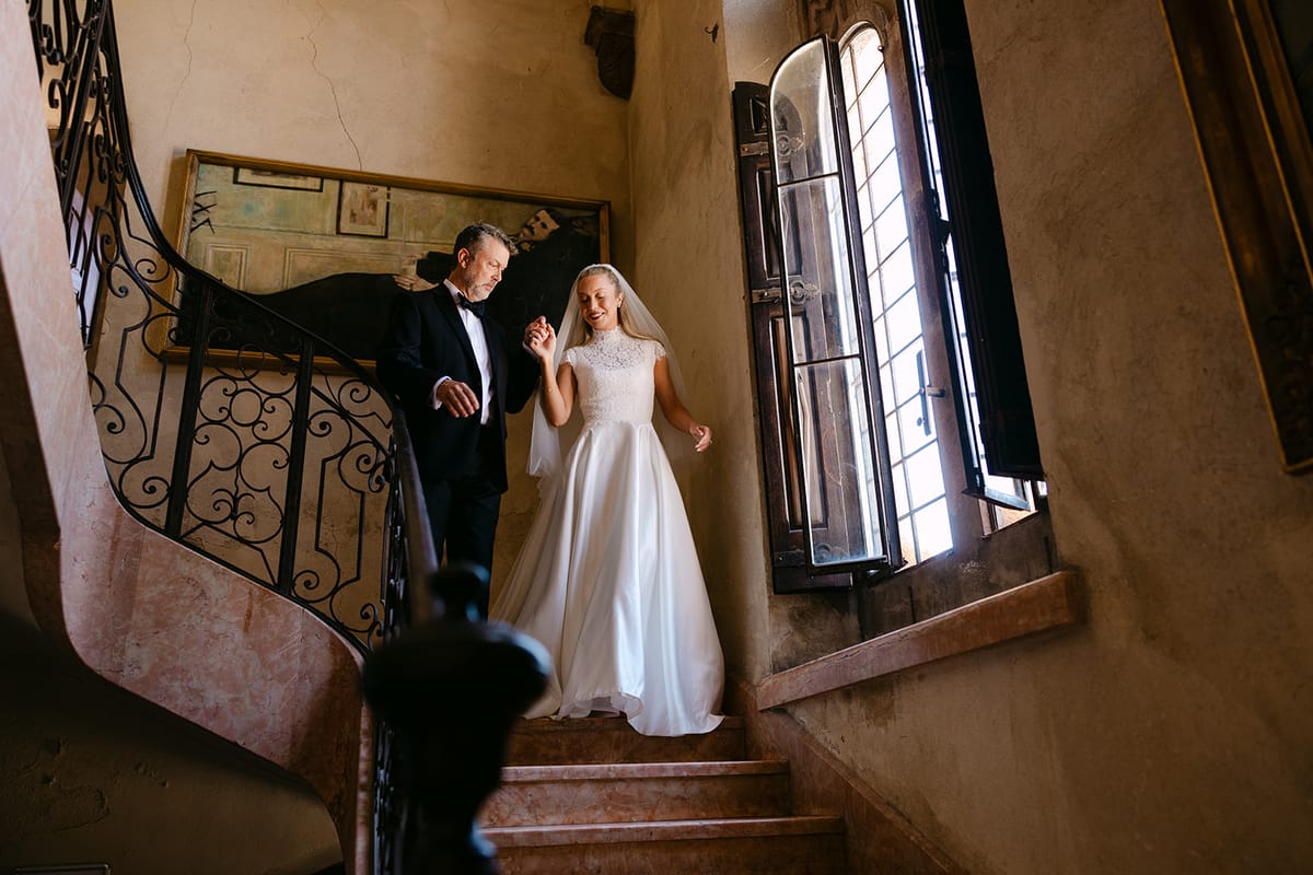 The bride in a white gown and veil walks down an old stone staircase beside a man in a dark suit.
