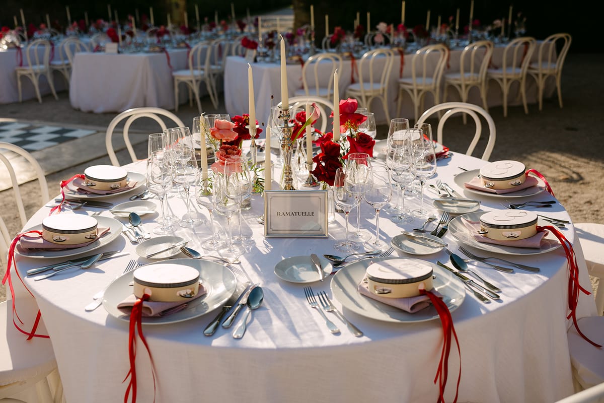 A round table laid with white plates, glasses, tall candles, red flowers and red ribbons, with more tables behind.
