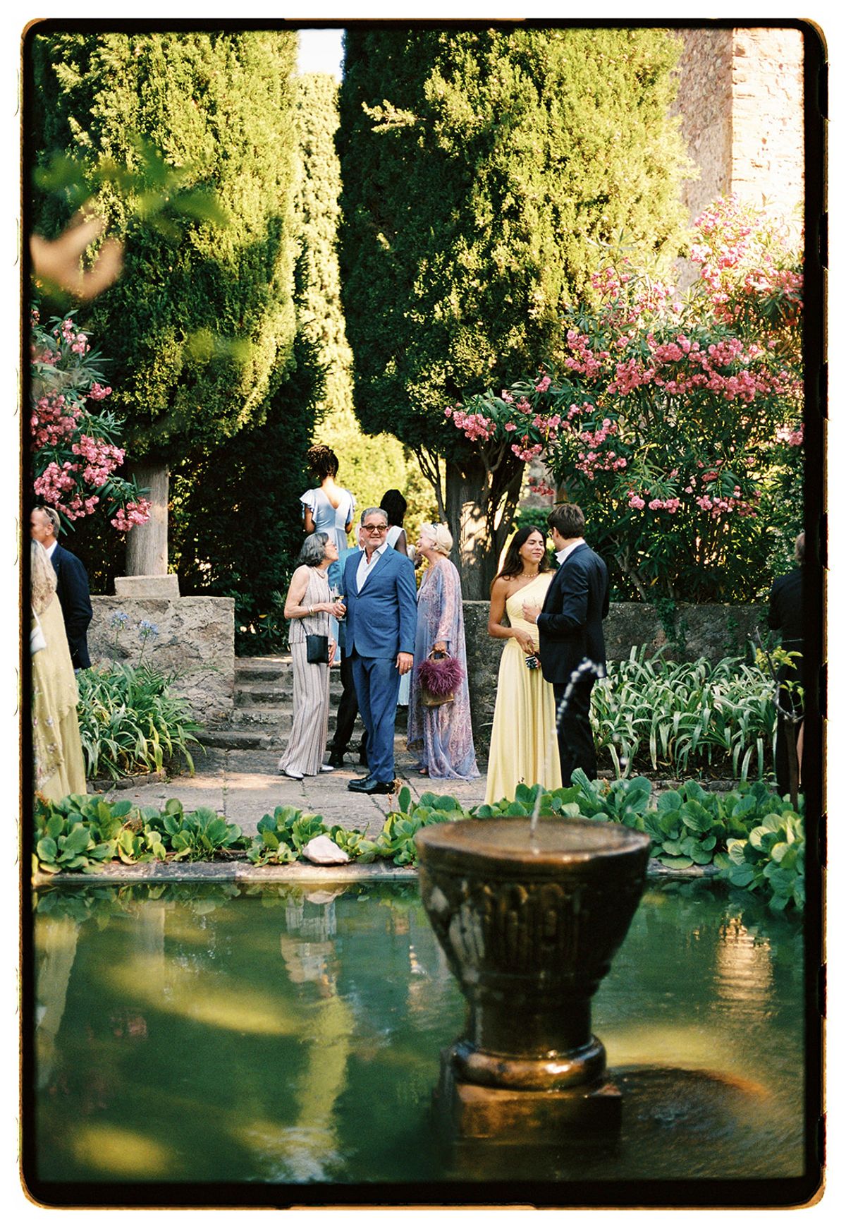 Guests in colourful outfits stand chatting in a lush garden behind a stone fountain and a still green pond.