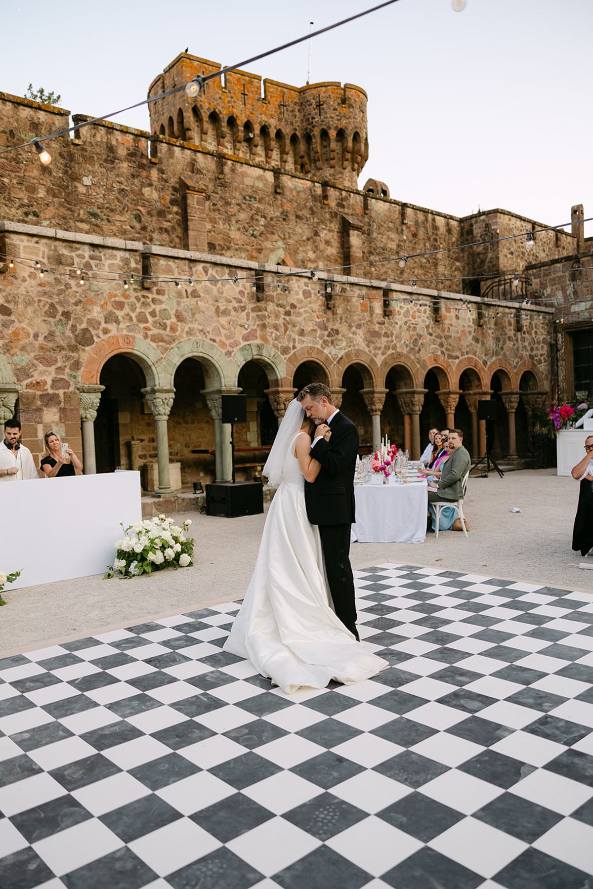 The bride and groom dance close on a chequered dance floor in front of a stone castle with arched cloisters and a crenellated tower.