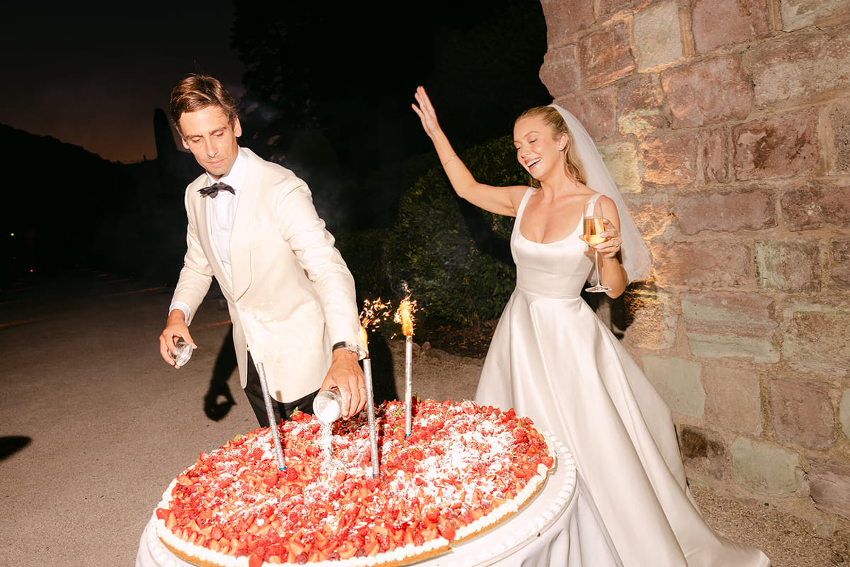 The groom and bride stand beside a large strawberry-topped cake with lit sparklers outside against a stone wall at dusk.