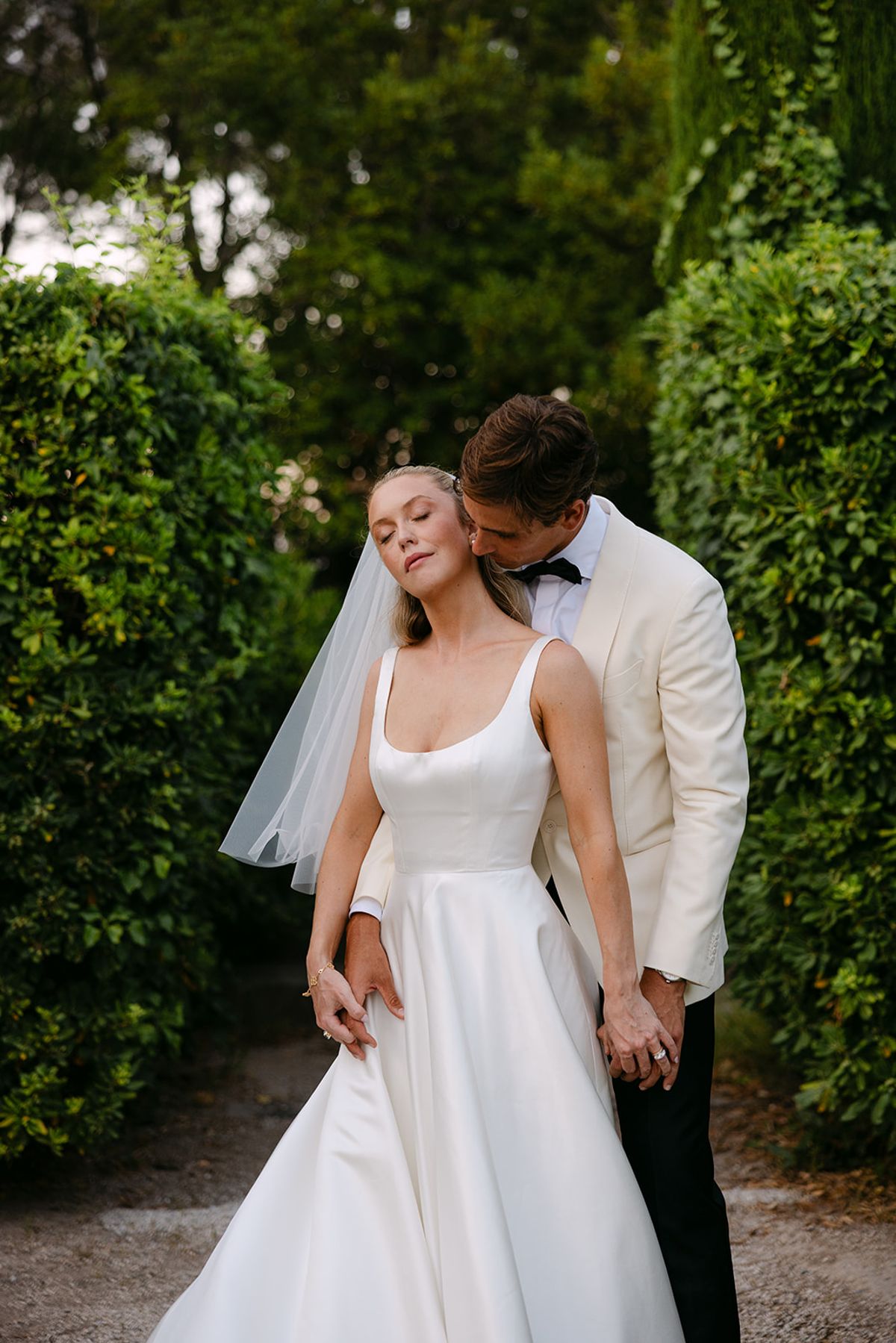 The groom in a white jacket nuzzles the bride from behind as she tilts her head back, with green hedges around them.