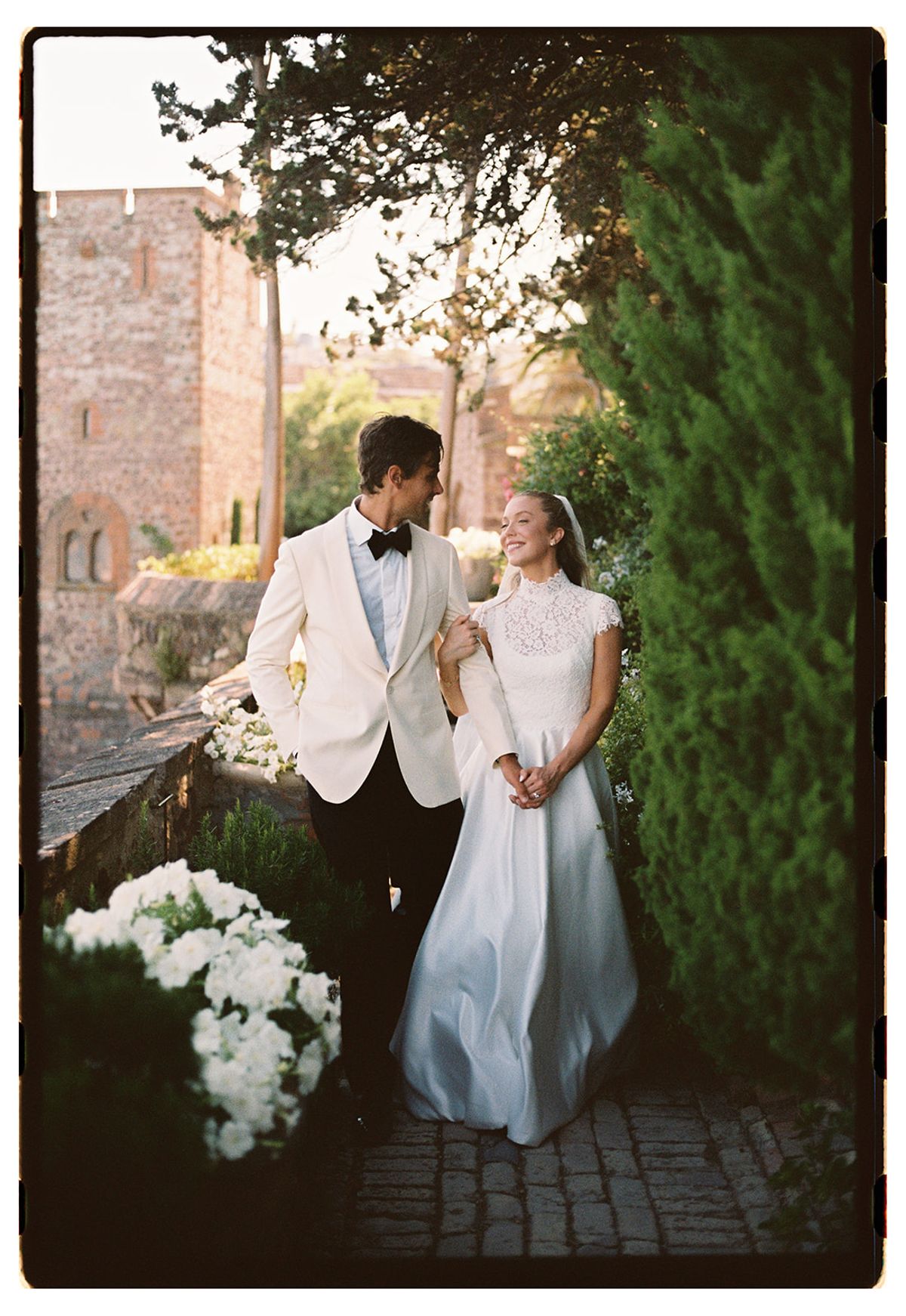 The couple stand close together, smiling at one another, beside white hydrangeas and a tall cypress tree near a stone wall.