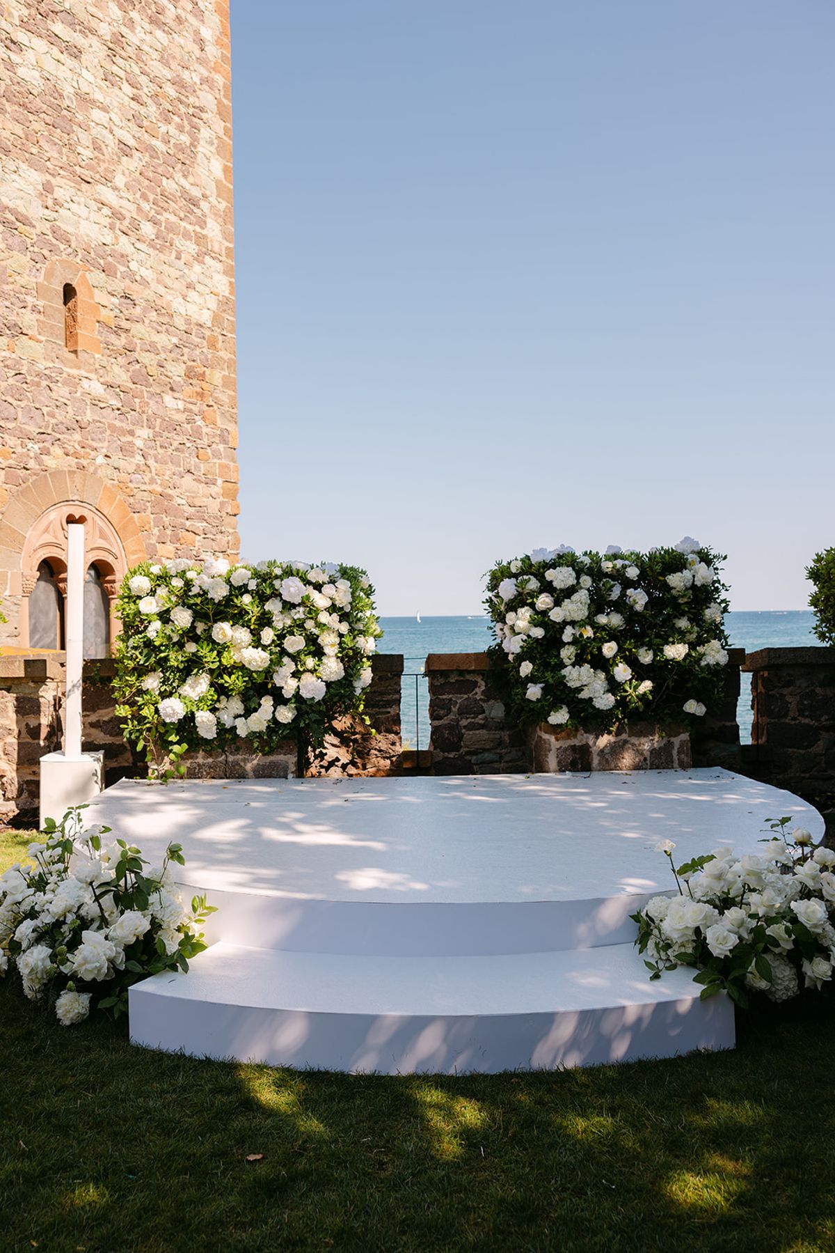 A white stepped platform flanked by white hydrangea arrangements stands on a lawn beside a stone tower, with the sea beyond.