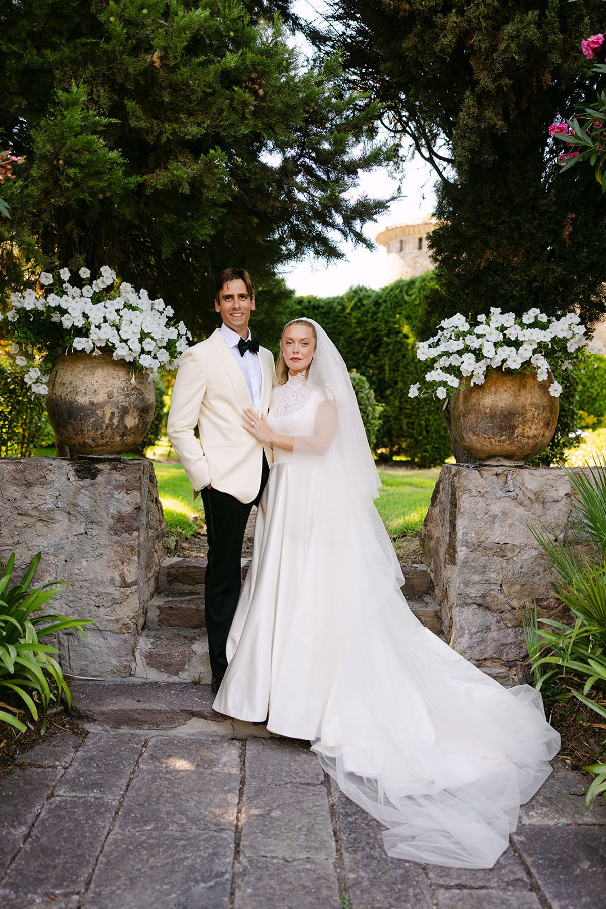The couple stand together on a stone path between urns of white flowers, the bride's veil and long train spread behind her.