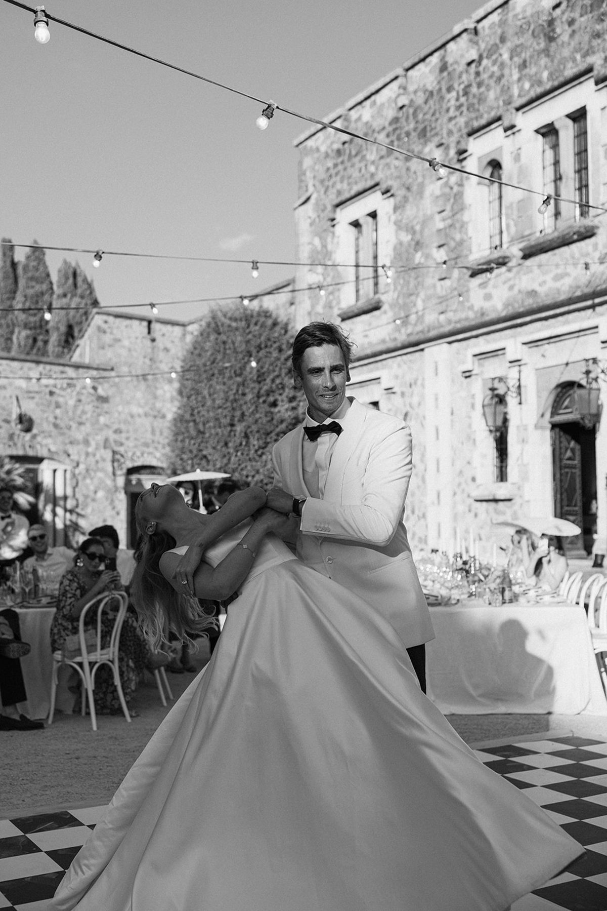 In black and white, the groom dips the bride, whose wide skirt sweeps across a checkered floor in a courtyard with string lights.