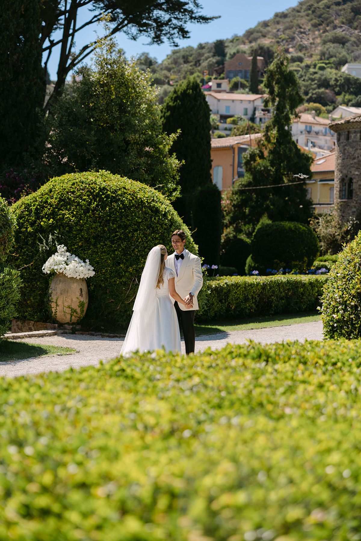 The bride in a veil and the groom in dark clothes stand close in a green garden, with a wooded hillside behind.