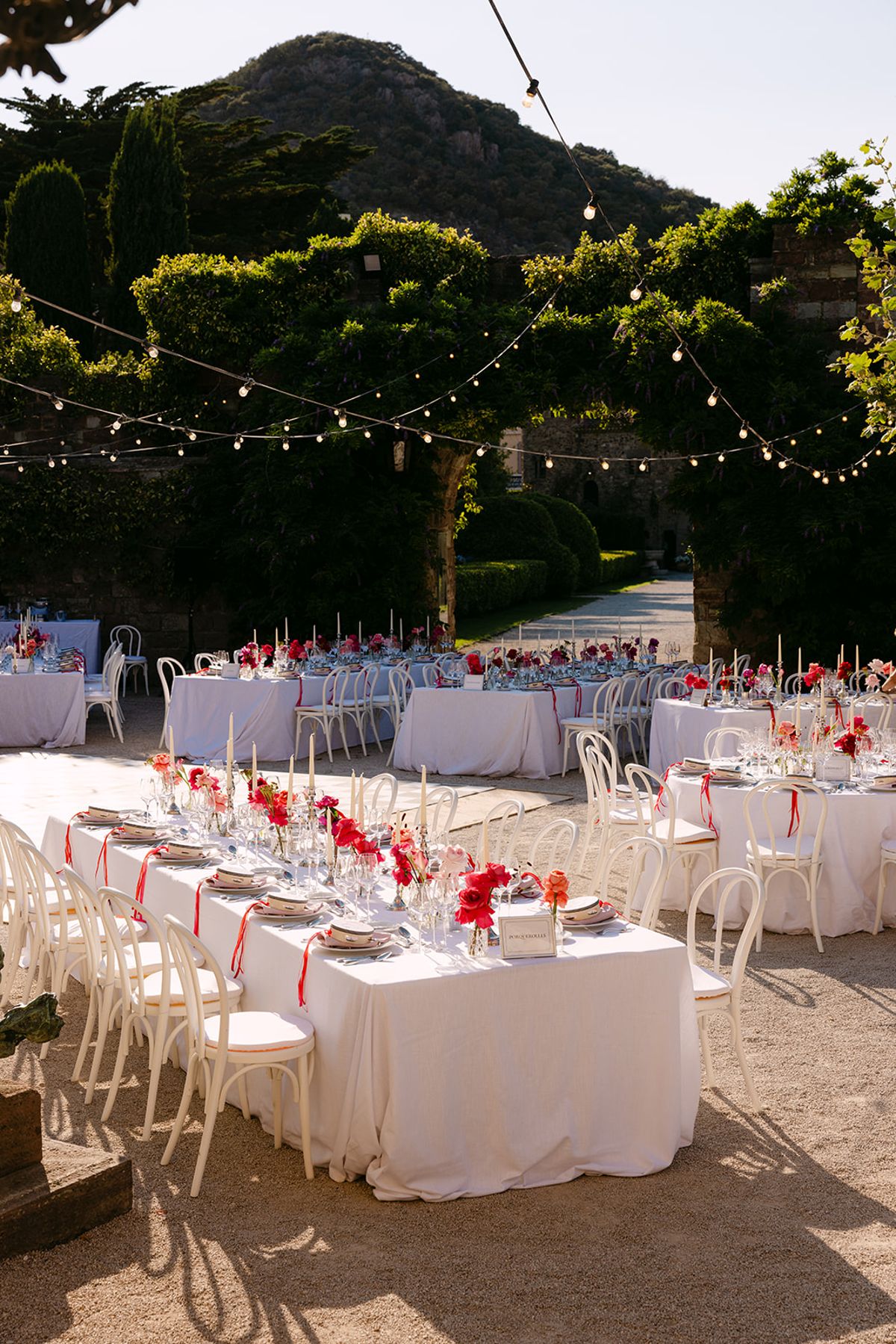 Long tables with white cloths and red floral centrepieces sit in a courtyard beneath string lights, with a hill behind.