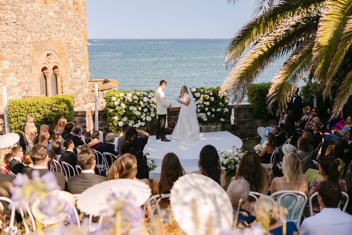 Guests seated facing the couple on a white platform surrounded by white flowers, with a stone tower and the sea behind.