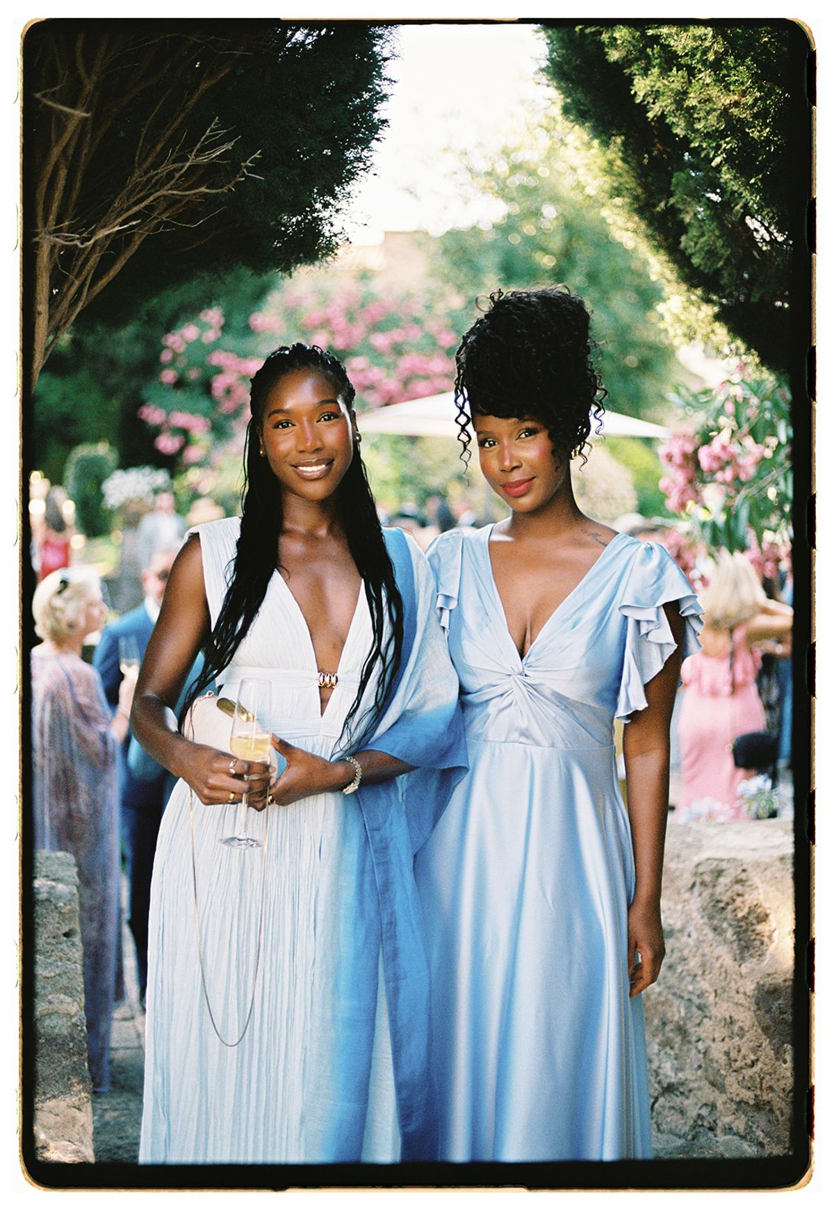 Two women stand side by side, one in a white dress and one in a pale blue dress, with guests behind them.