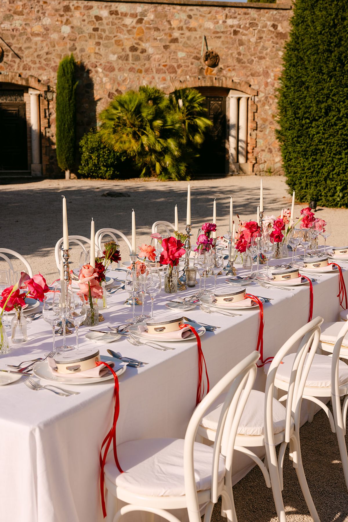 A long table with white linen, tall candles and red flowers, with white chairs tied with red ribbons in a stone courtyard.