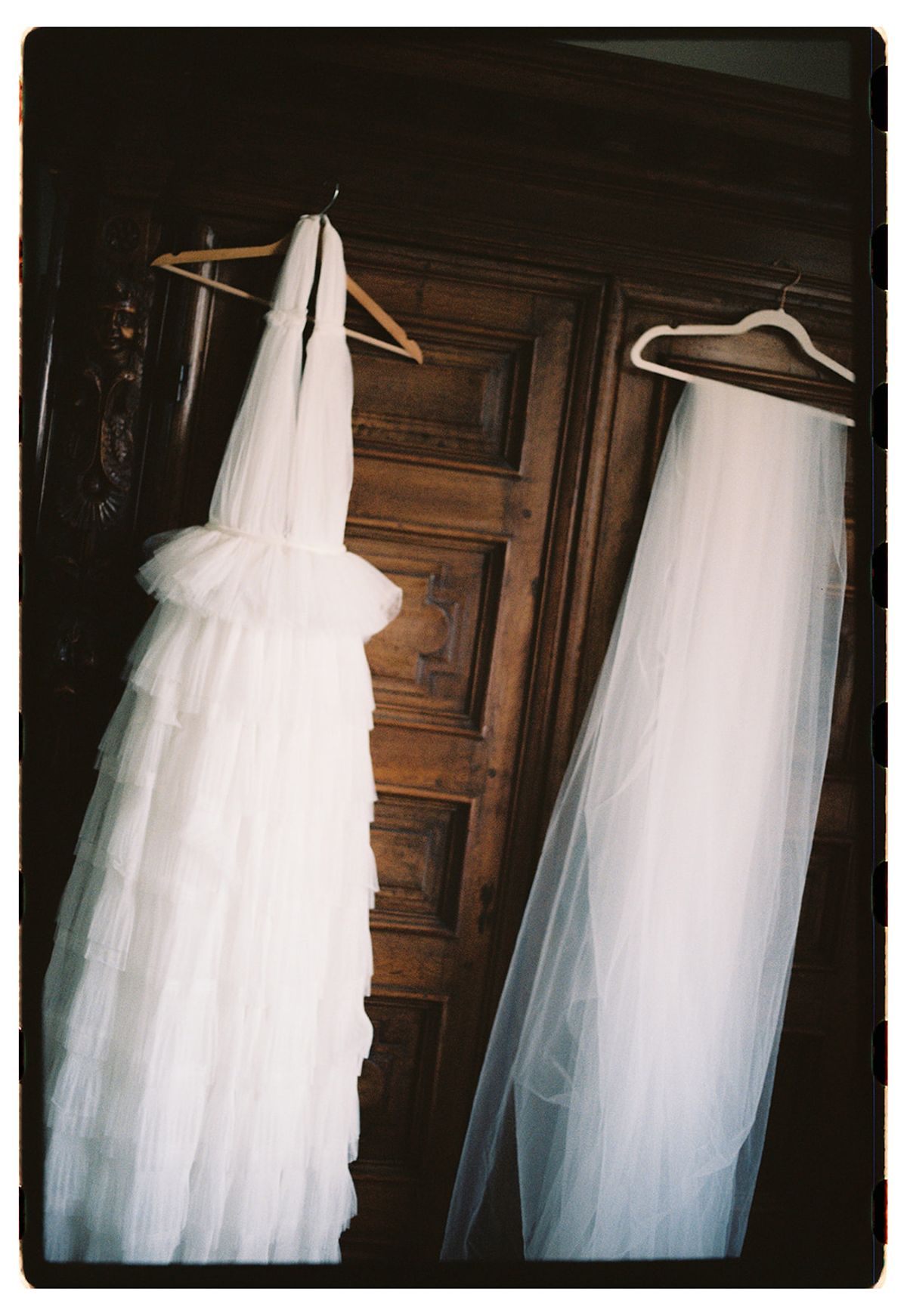 A tulle wedding dress and a long veil hang from hangers against a dark panelled wooden door.