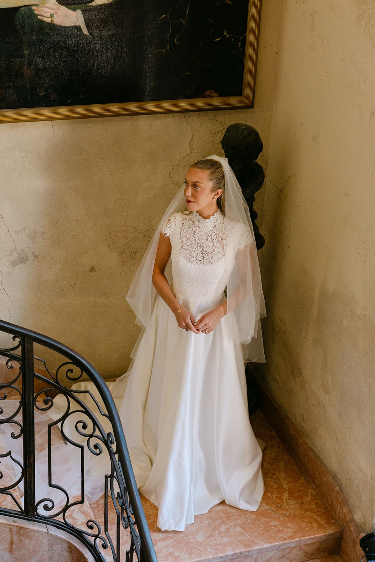 The bride in a high-necked gown and veil stands smiling beside a wrought-iron stair balustrade in a warm-toned hallway.