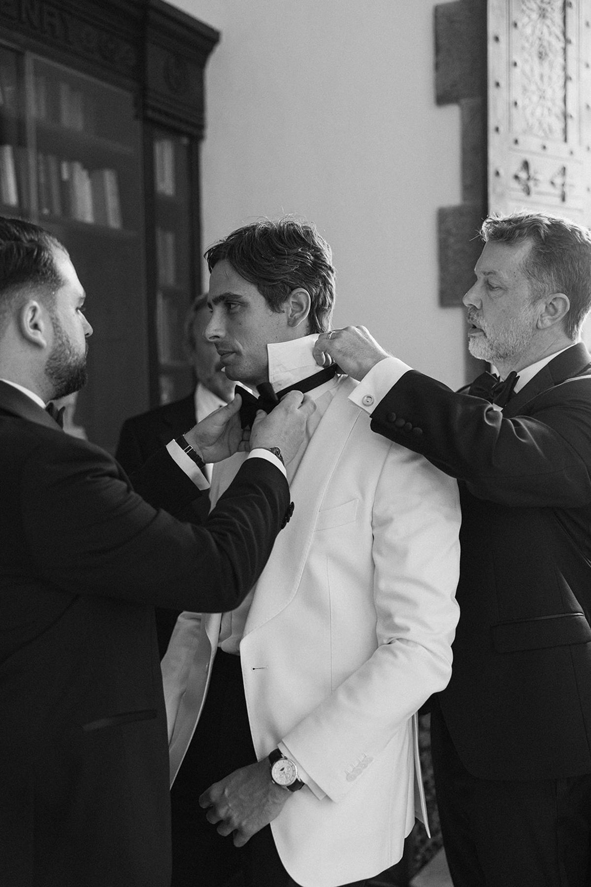 In black and white, two men in dark suits help the groom in a white jacket adjust his bow tie.