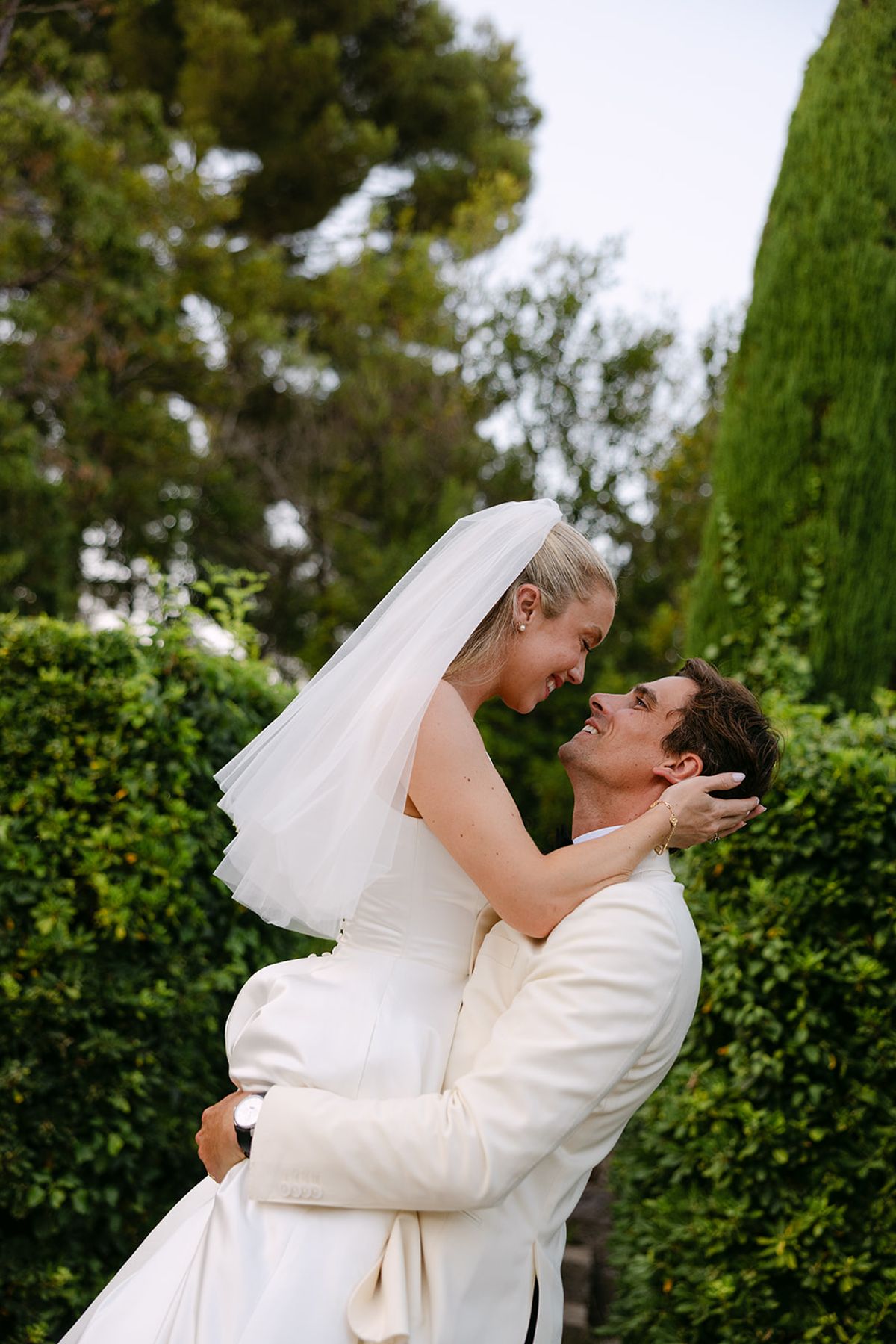 The groom lifts the veiled bride in an embrace as they lean in to kiss, with tall green hedges behind.