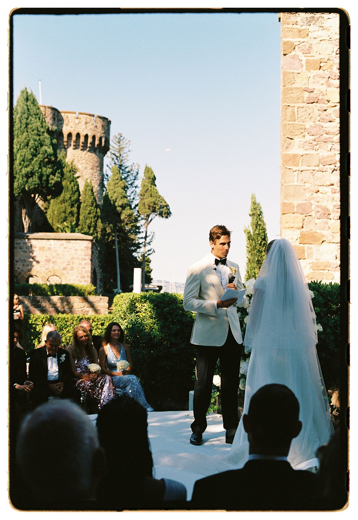 A man in a light jacket speaks beside the veiled bride in front of seated guests, with a stone tower behind.