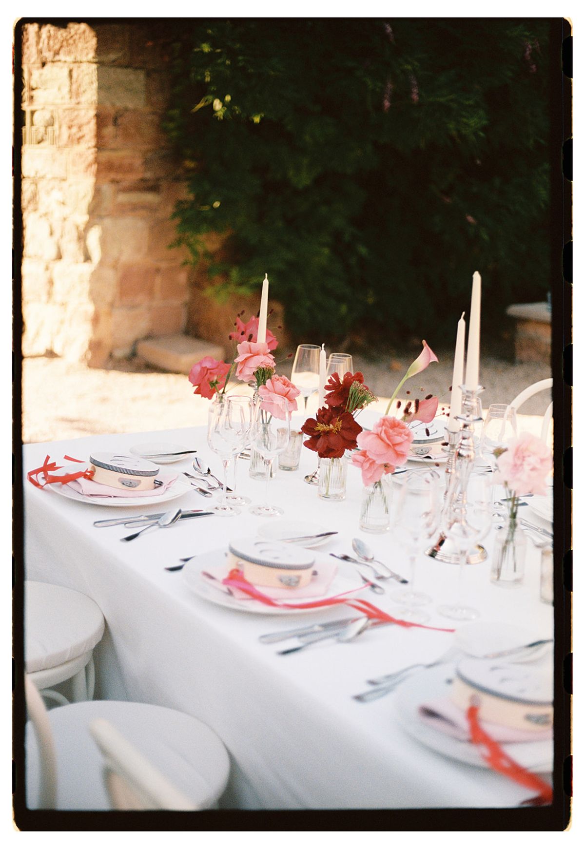 A long table with a white cloth is set with plates tied with red ribbon, candles and small vases of pink and red flowers.