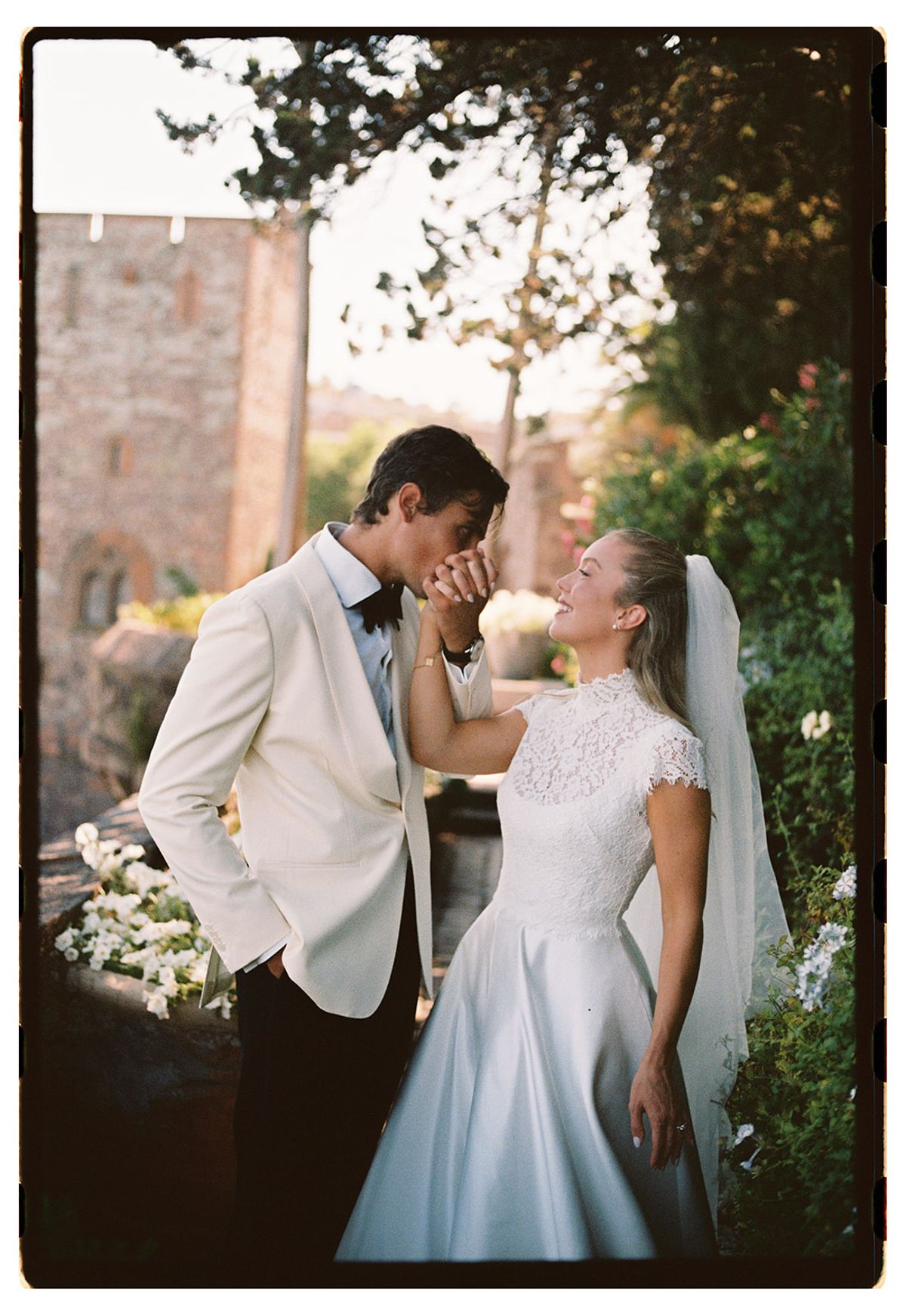 The groom in a white jacket kisses the bride's hand while she smiles, with a stone tower and greenery behind.