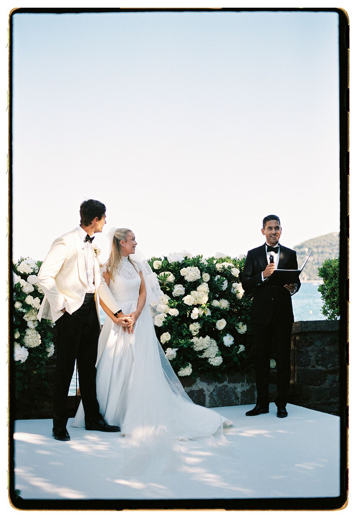 The couple stand on a white platform before white hydrangeas while an officiant in dark clothes reads beside them.