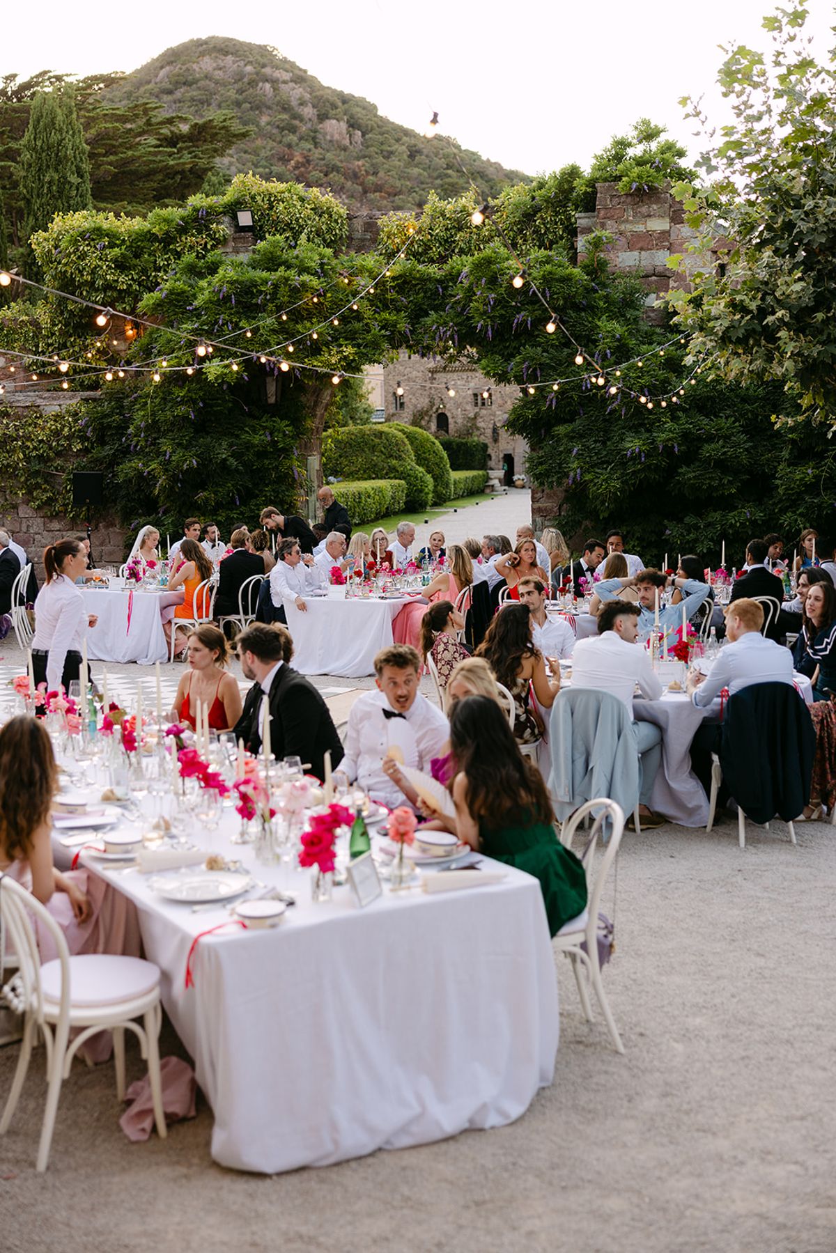 Guests sit at long white-clothed tables in a garden courtyard beneath strings of lights, with hills in the distance.