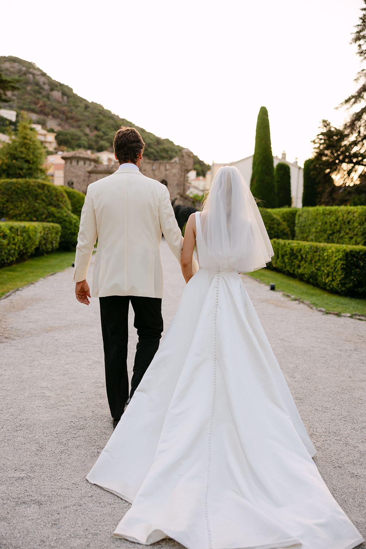 Seen from behind, the groom in a white jacket and the bride in a long gown and veil walk along a gravel garden path.