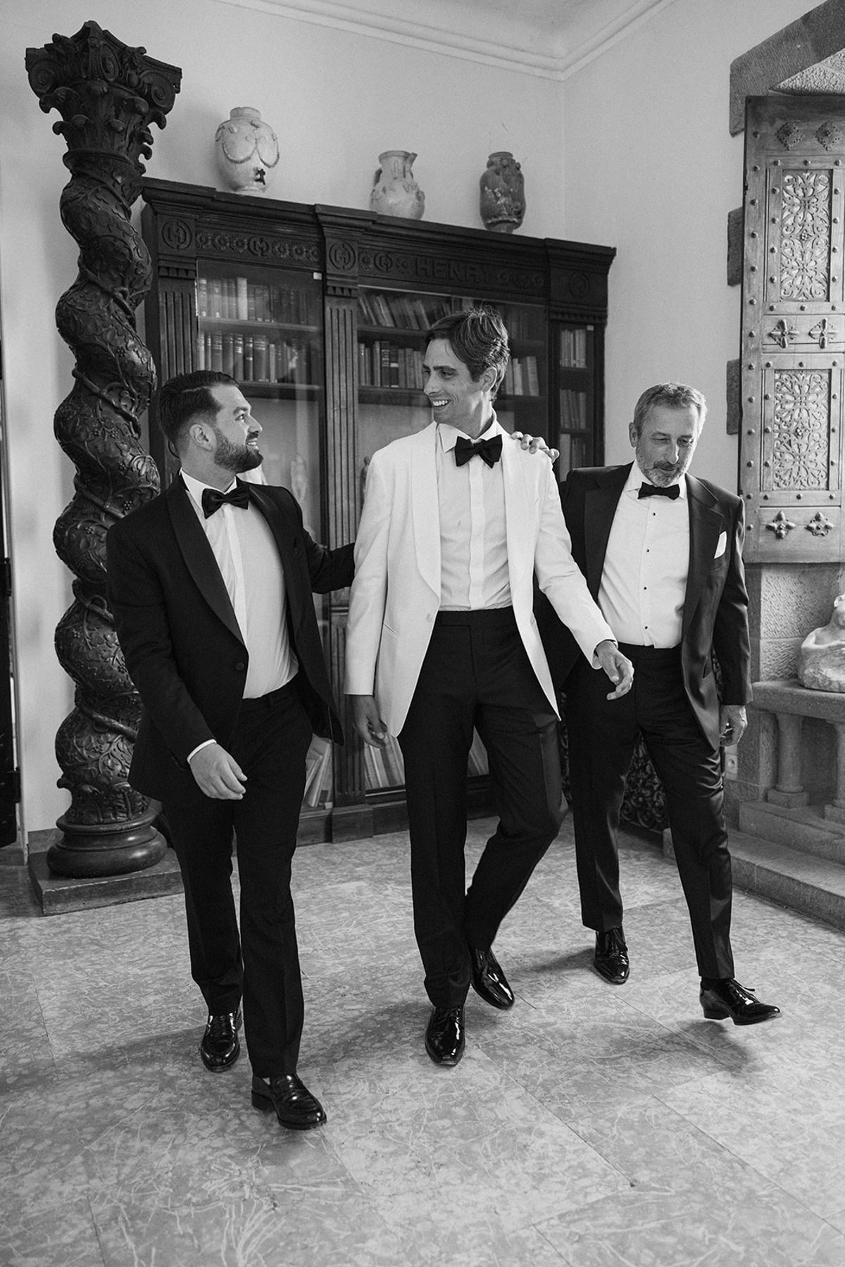 In black and white, the groom in a white jacket walks between two men in dark suits in a room with bookcases.