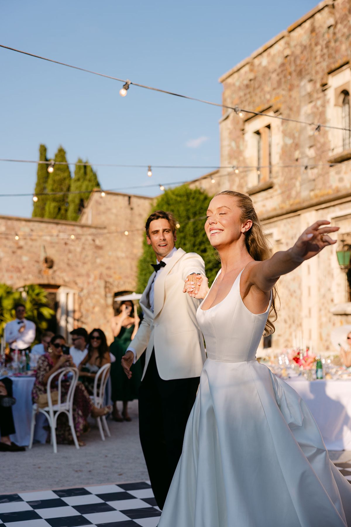 The bride and groom walk into the courtyard dinner as the bride reaches out an arm, with seated guests and festoon lights behind.
