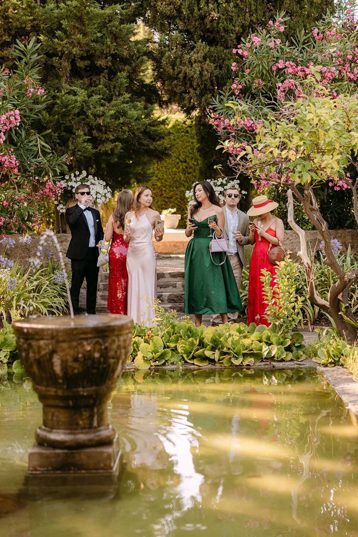 A man and several women in red and green dresses stand together beside a stone fountain pond among flowering trees.