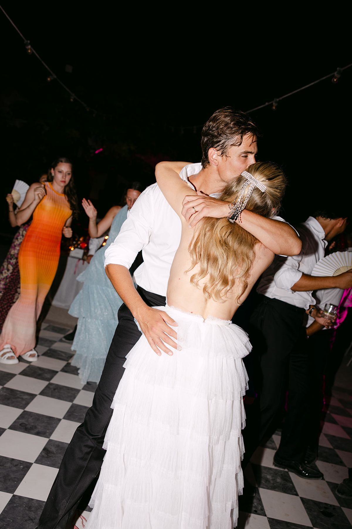The bride in a white tiered gown and the groom embrace on a checkerboard dance floor while guests dance around them.
