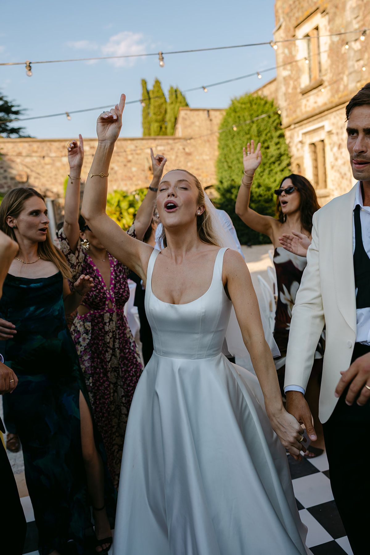The bride in a white dress sings with her arms moving while guests around her dance and raise their hands.