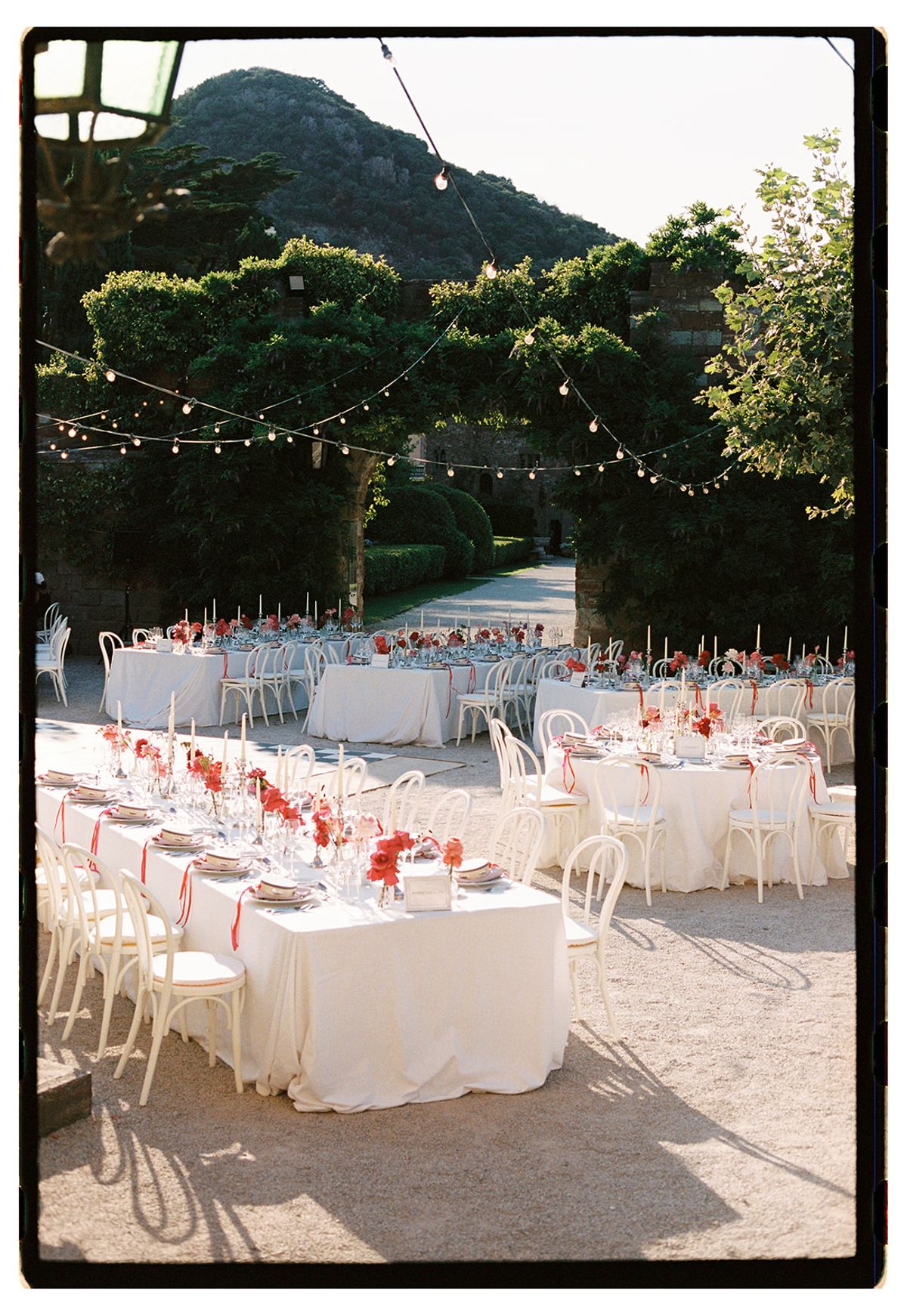 Long white tables with white bentwood chairs sit on gravel beneath trees and strung lights, with a hill in the distance.