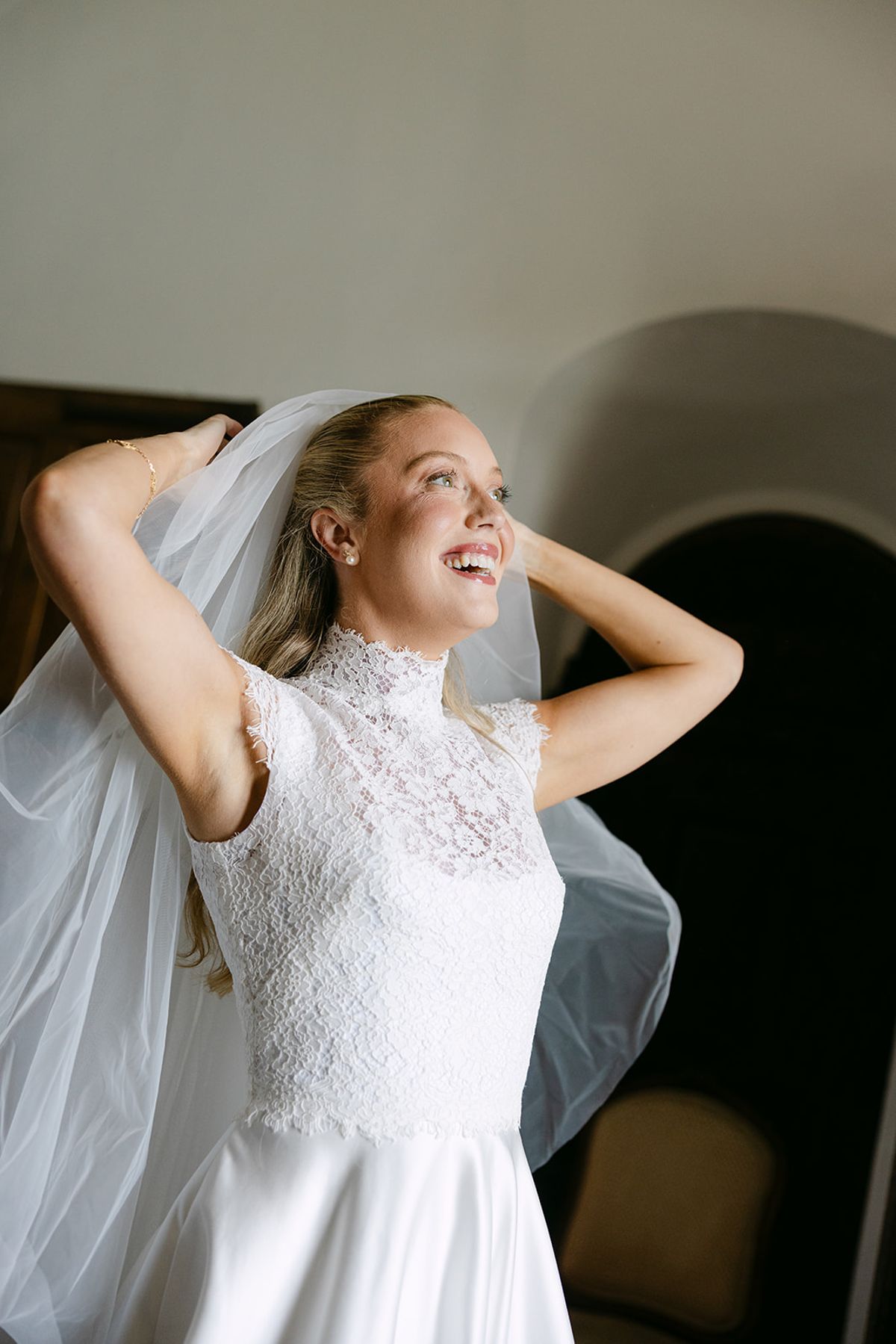 The bride in a high-necked lace bodice looks up and smiles as she adjusts her veil with both hands.