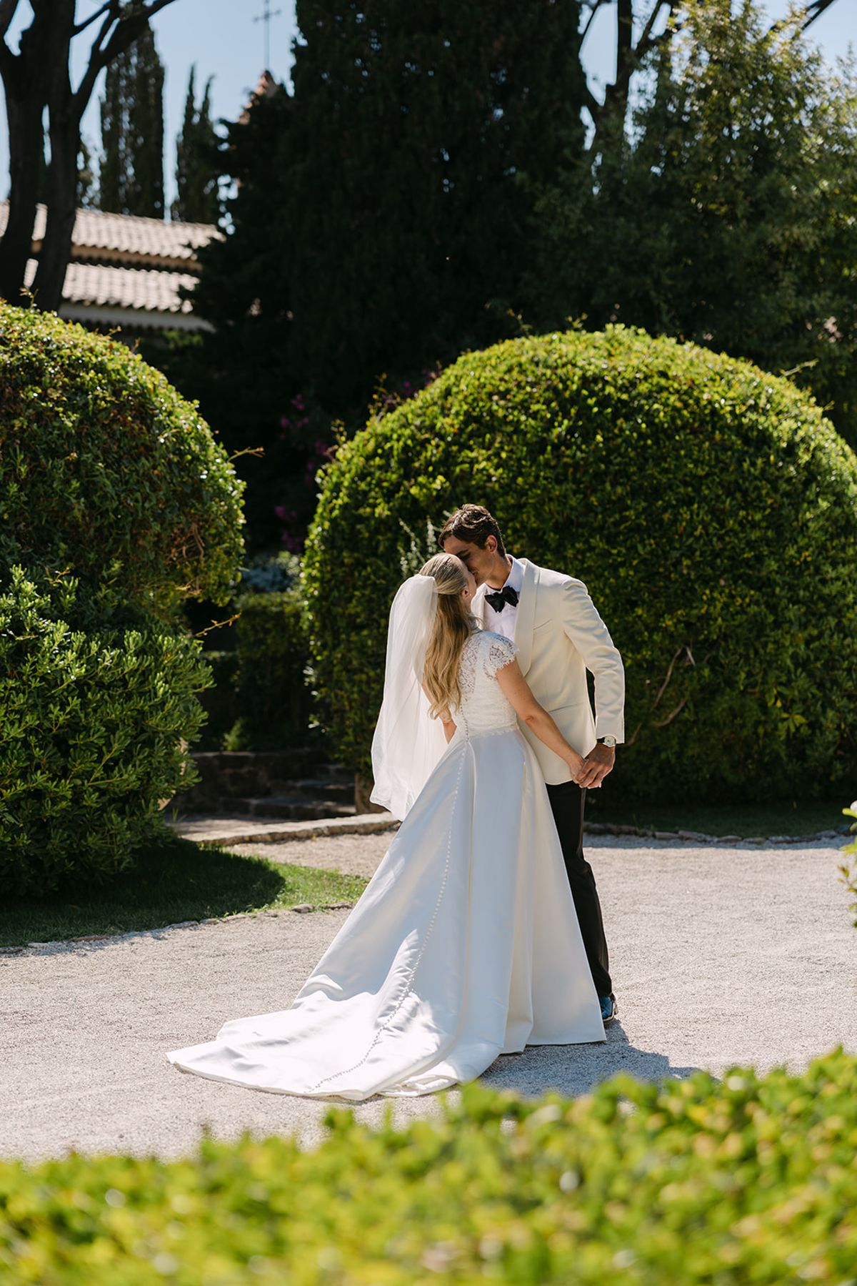 The couple embrace on a gravel path in front of rounded green topiary, the bride's long veil and dress trailing behind.