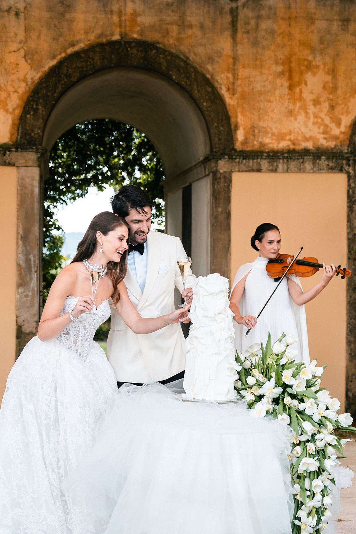 Bride and groom cutting the ruffled white cake as a violinist plays beneath the arcade at Château de Sannes, Provence