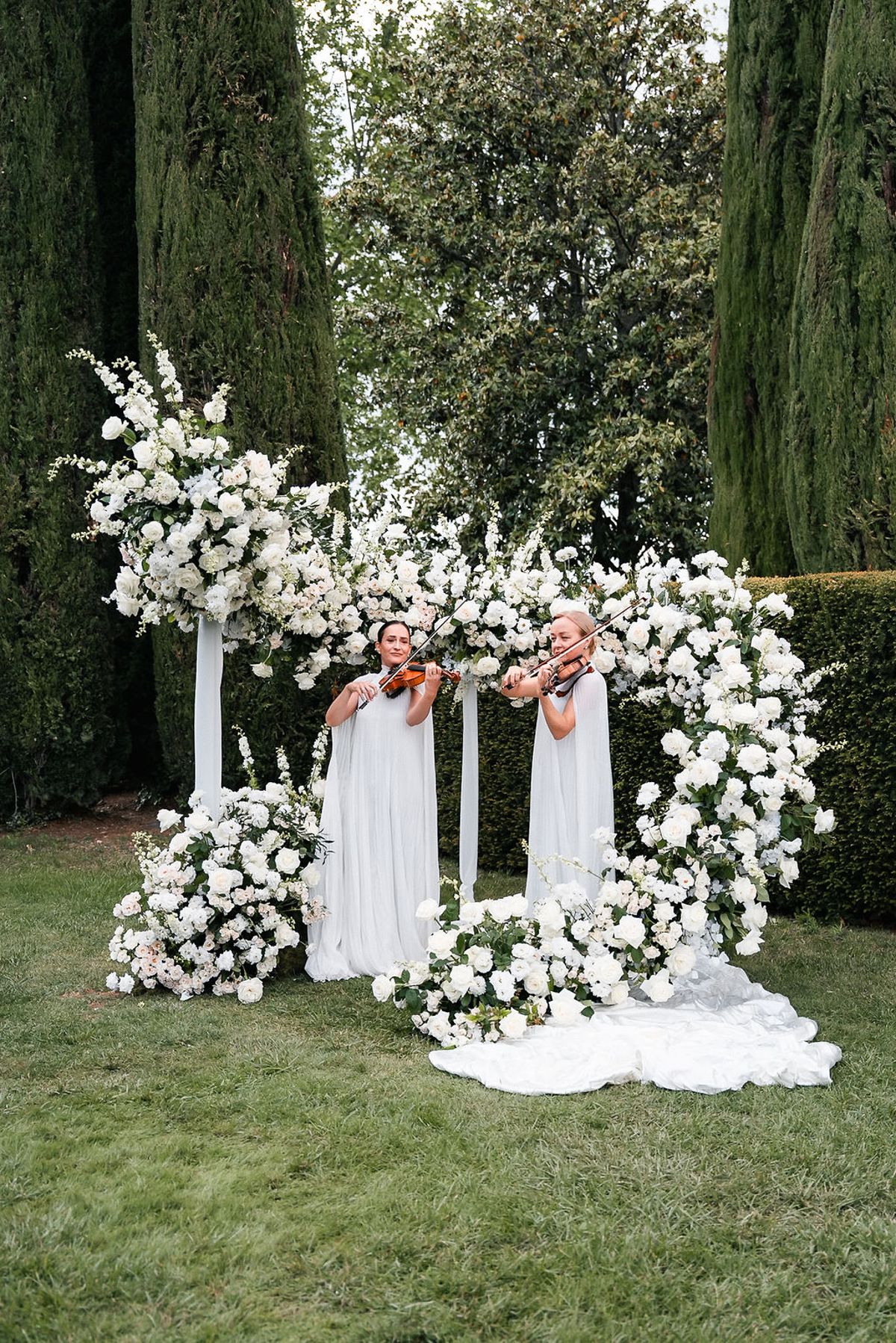 Two violinists in white playing beside a white floral installation at the ceremony setting at Château de Sannes, Provence