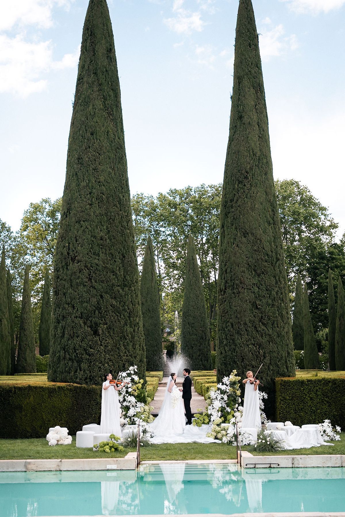 Ceremony framed by two tall cypresses across the swimming pool, with violinists either side at Château de Sannes, Provence