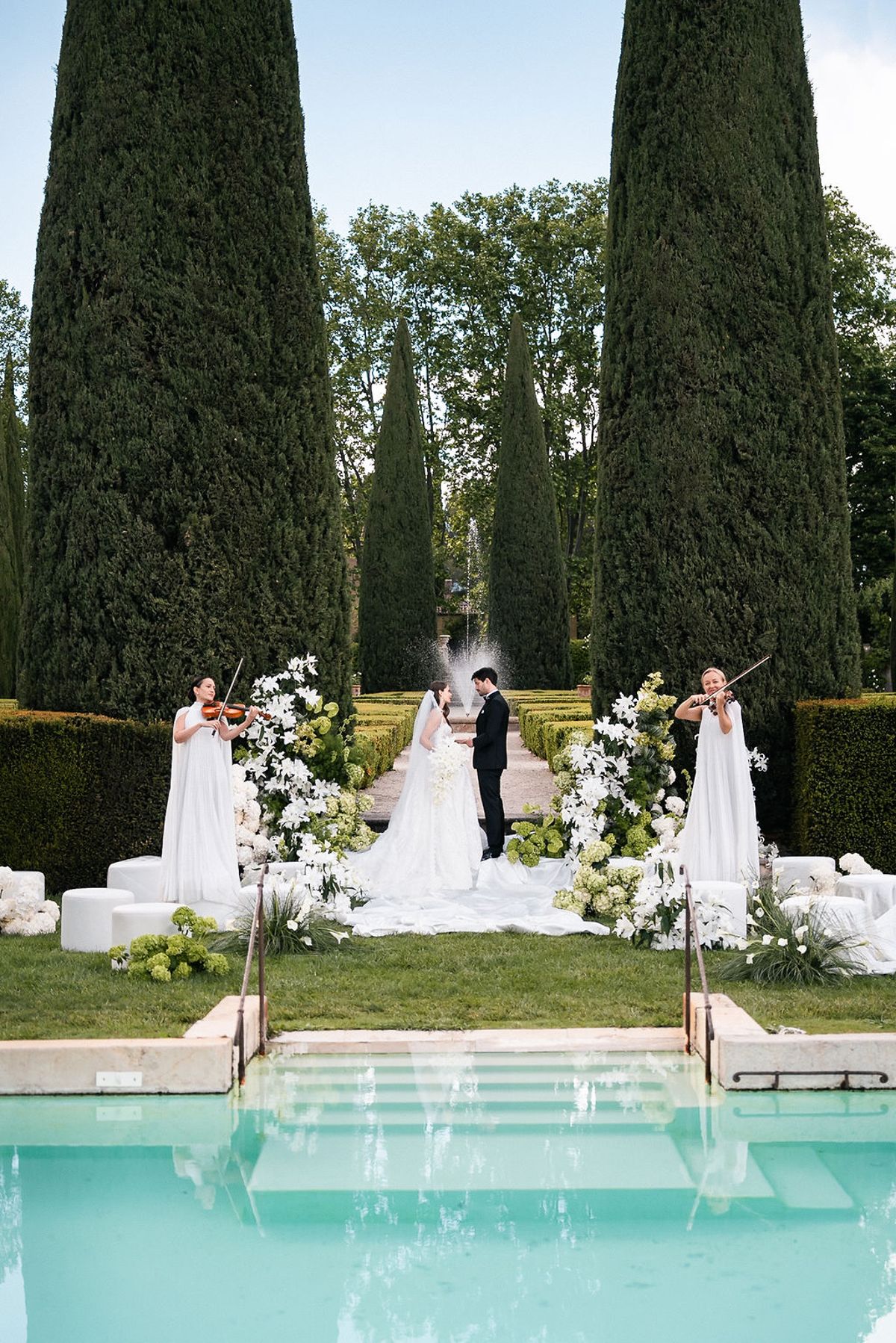 Ceremony staged on the lawn beyond the swimming pool, framed by white florals and cypresses at Château de Sannes, Provence
