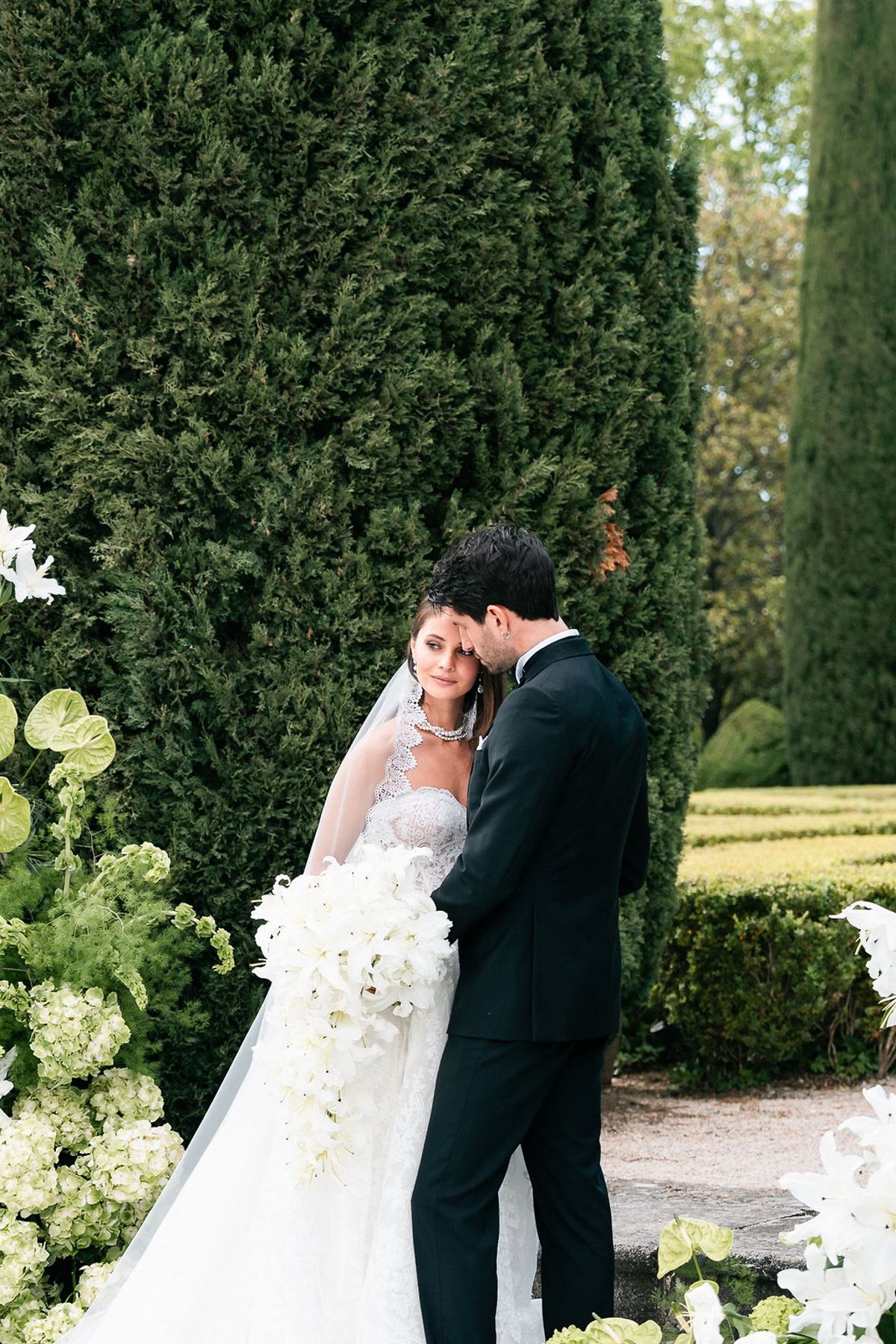 Bride holding a cascading white bouquet against a groom in a black tuxedo before a wall of greenery at Château de Sannes, Provence