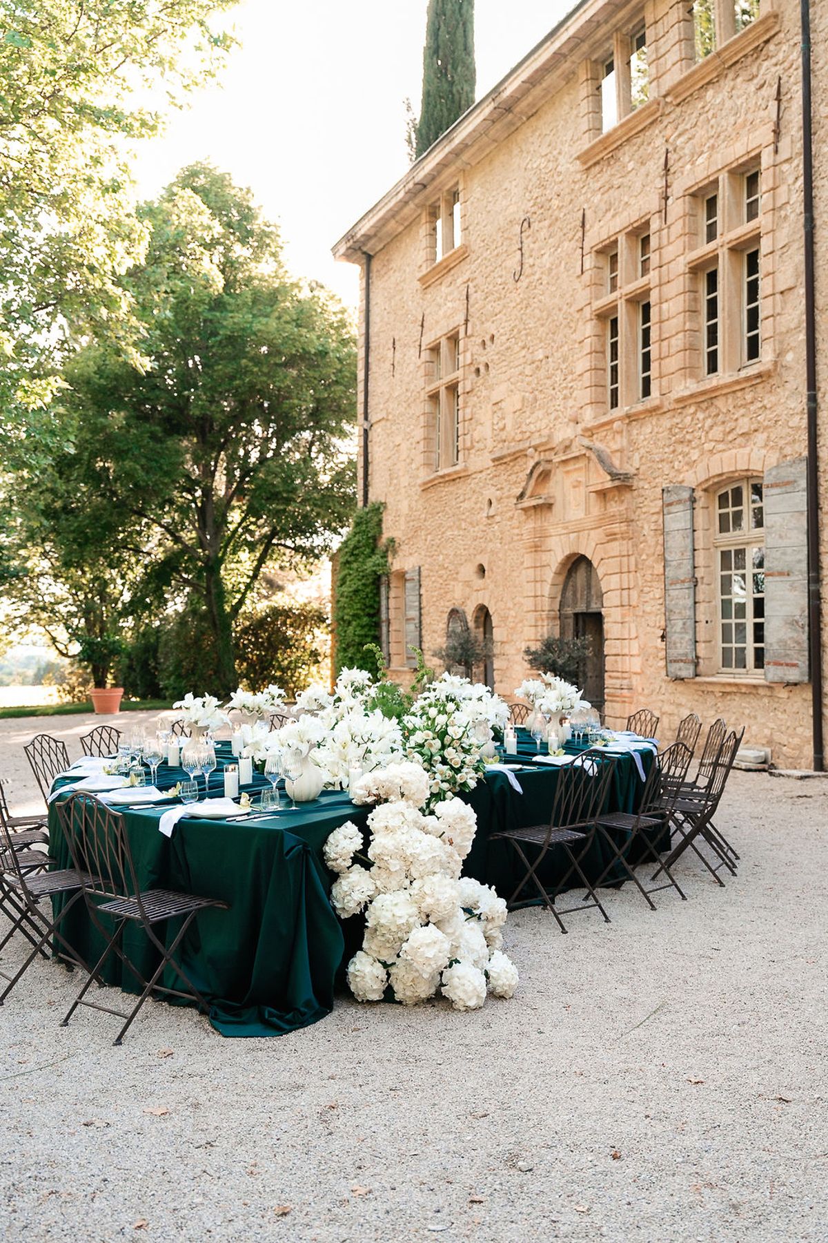 Green-dressed dining tables and white florals arranged on the gravel below the château at Château de Sannes, Provence