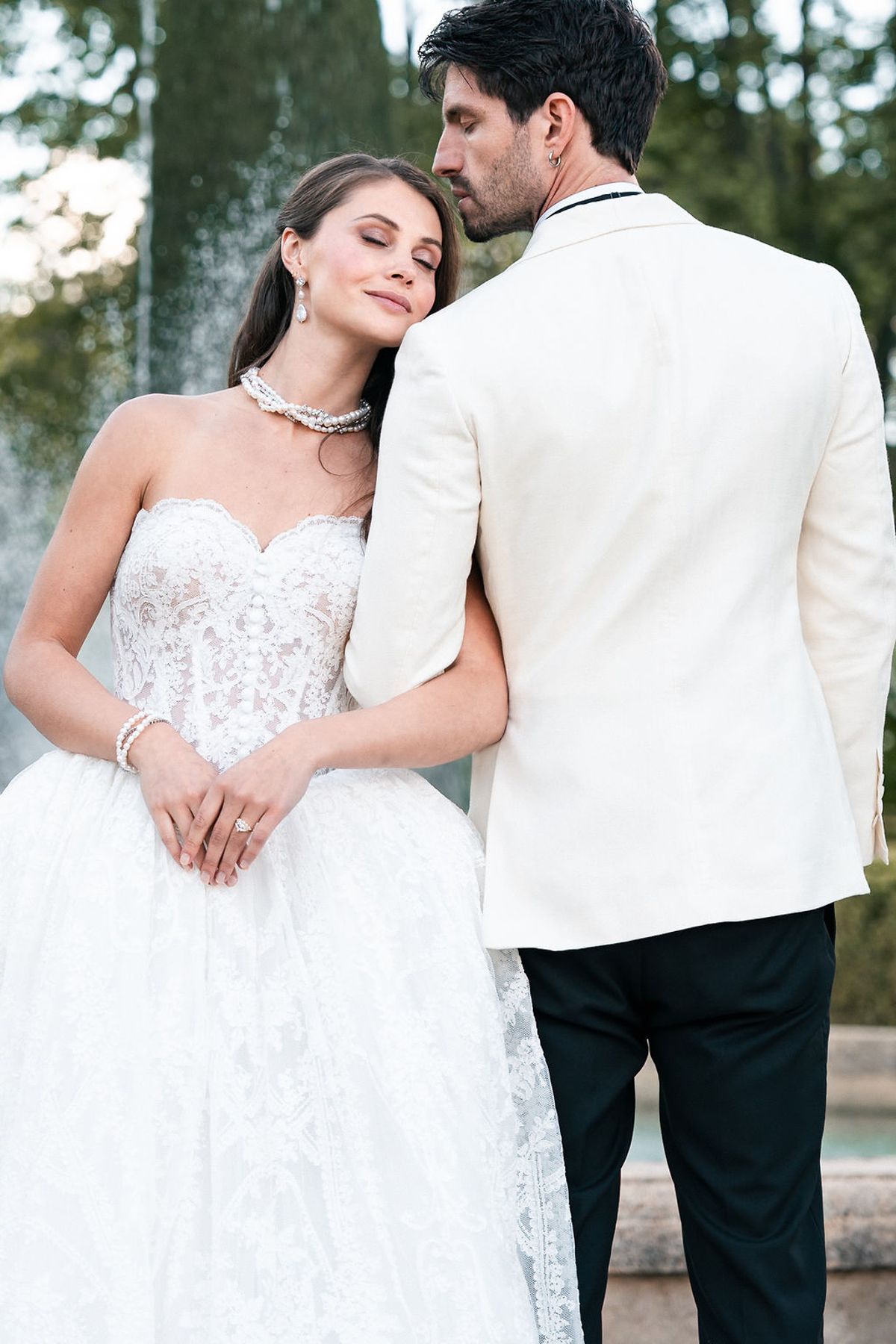 Bride in a strapless lace ball gown resting against a groom in a white dinner jacket in the gardens at Château de Sannes, Provence