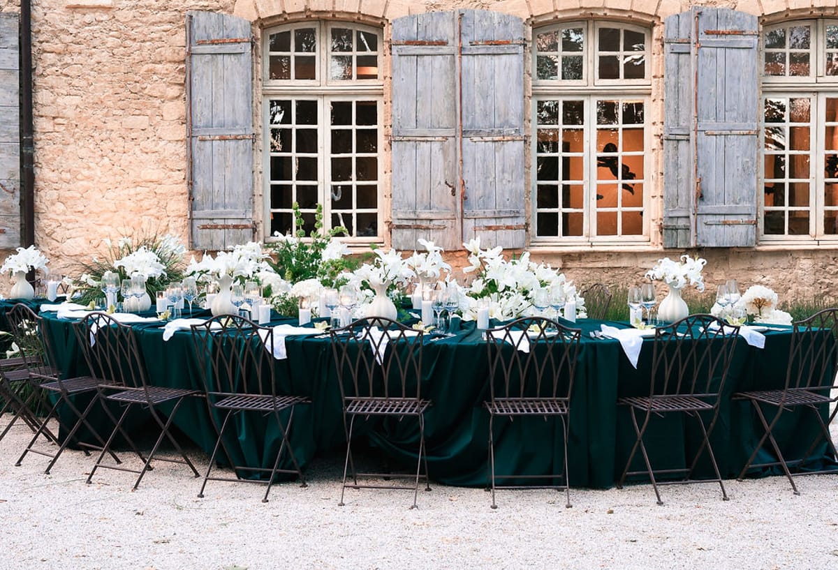 Two long green-dressed tables set with white flowers below the shuttered stone façade at Château de Sannes, Provence