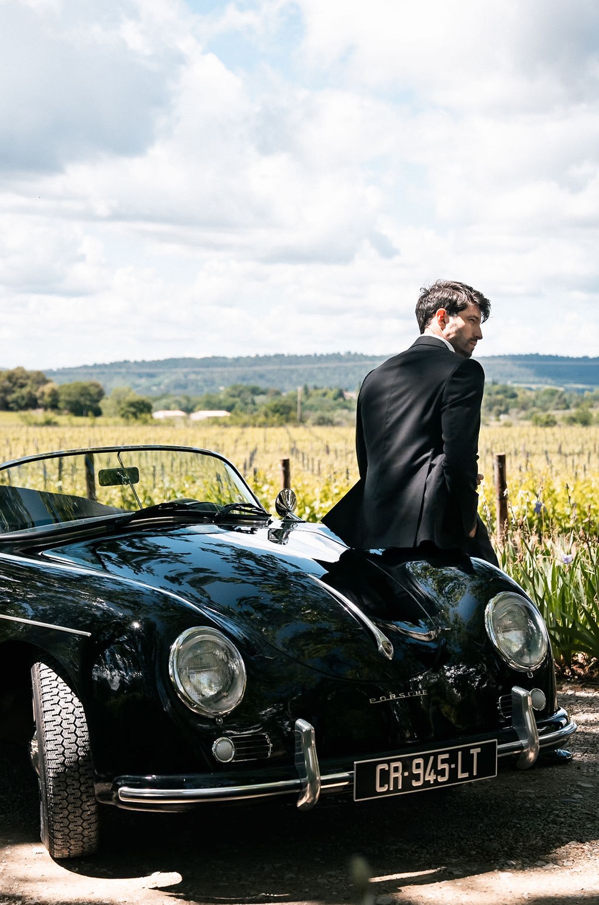 Groom in a black tuxedo seated on the bonnet of a vintage open-top Porsche beside the vines at Château de Sannes, Provence