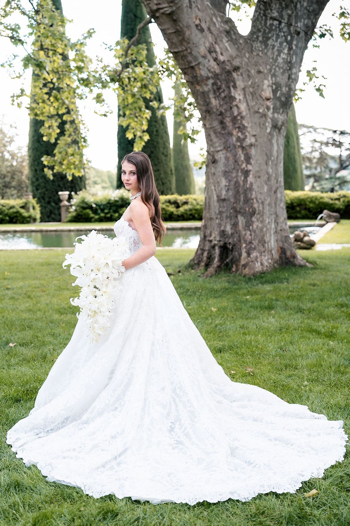 Bride holding a cascading white lily bouquet beneath an old plane tree on the lawn at Château de Sannes, Provence