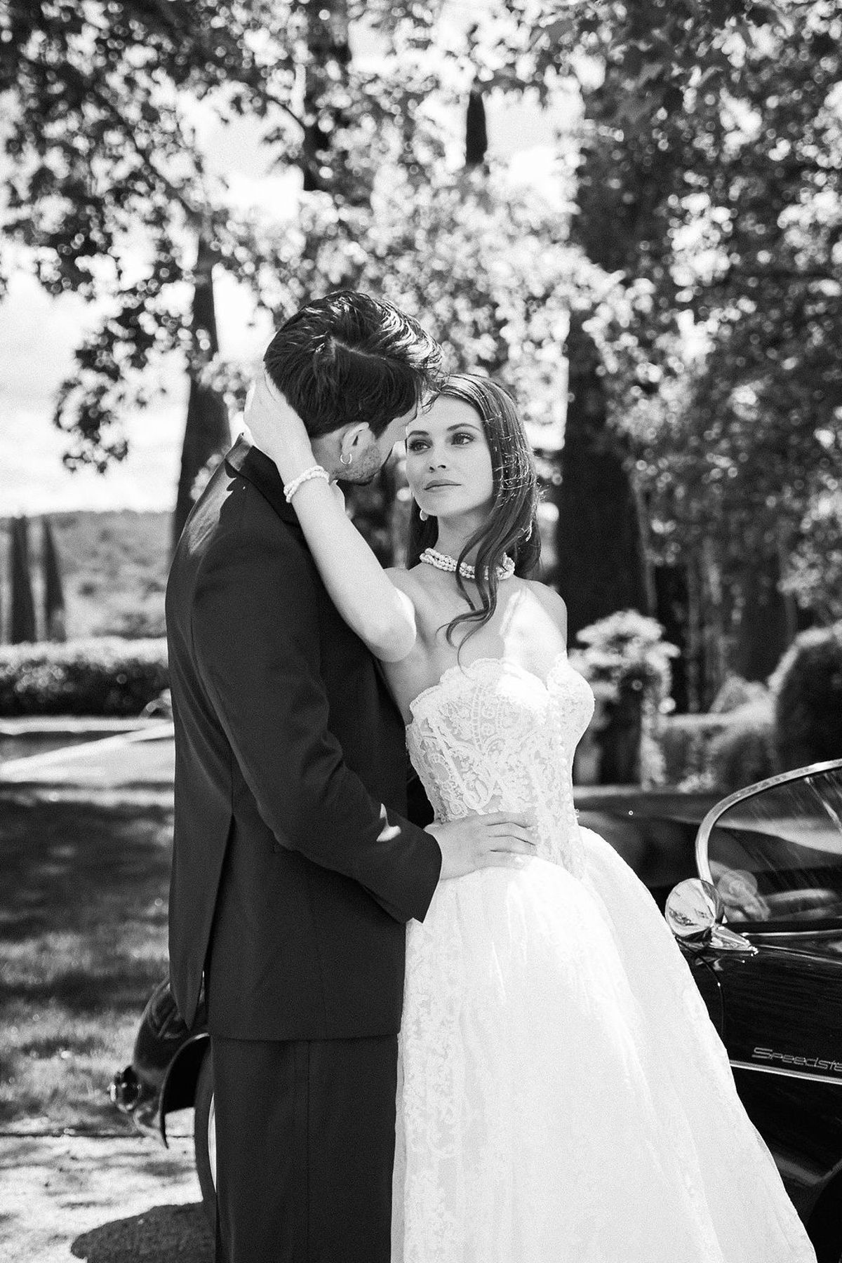 Black and white portrait of a bride and groom embracing beside a vintage open-top car at Château de Sannes, Provence