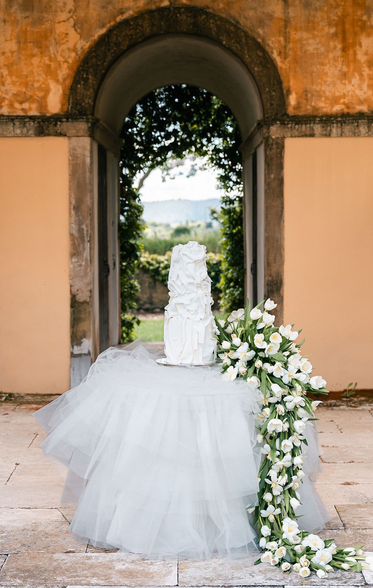 Tiered white ruffled wedding cake on a tulle-draped table under the ochre-rendered arcade at Château de Sannes, Provence