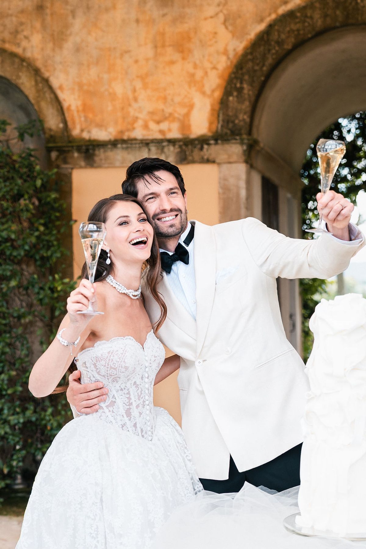Bride and groom raising champagne flutes beneath an ochre-rendered arcade hung with ivy at Château de Sannes, Provence