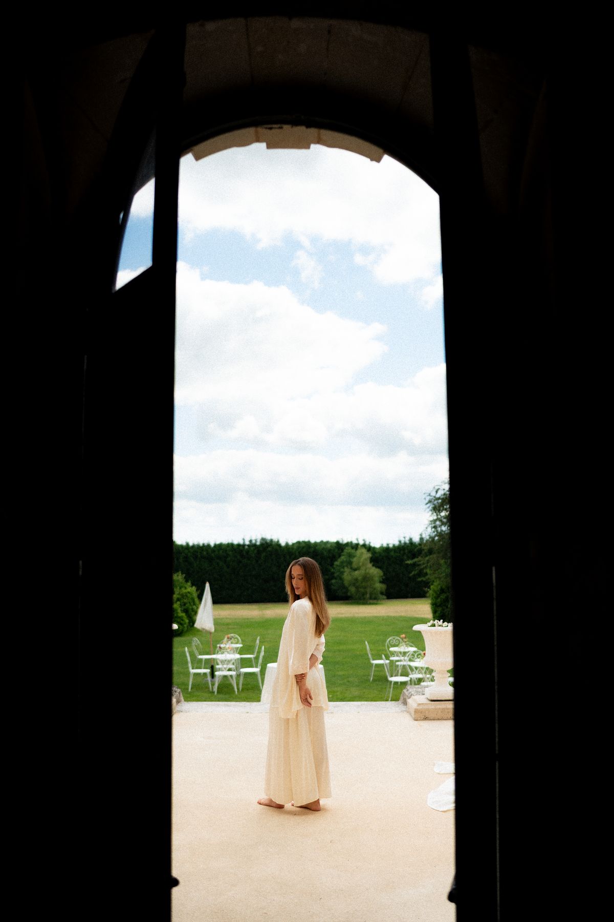 Isadora & Hugo — the bride framed by a stone arch with the lawn beyond, spring wedding in Nouvelle-Aquitaine