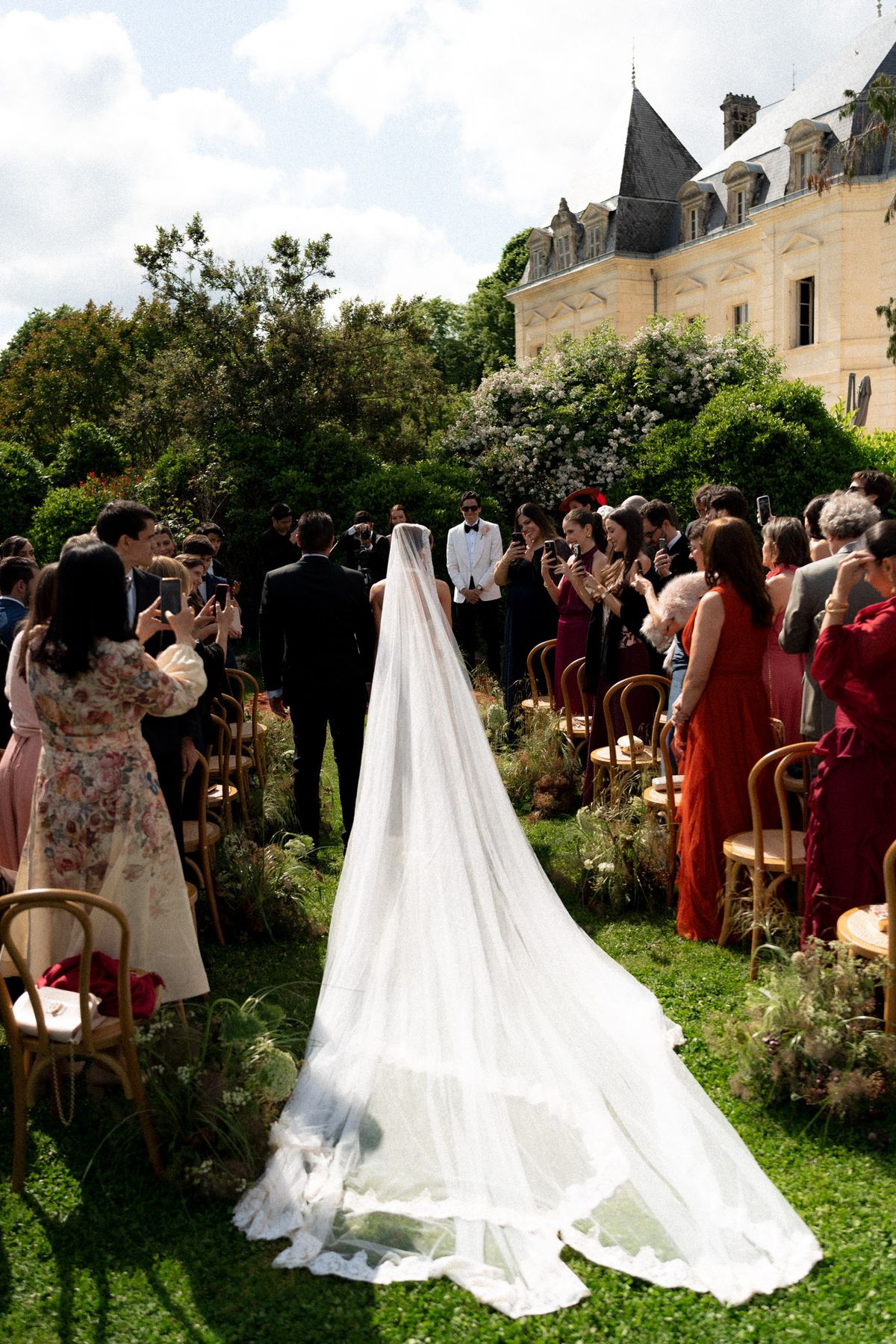 Isadora & Hugo — the bride from behind, her long veil trailing across the grass toward the seated guests, spring wedding in Nouvelle-Aquitaine